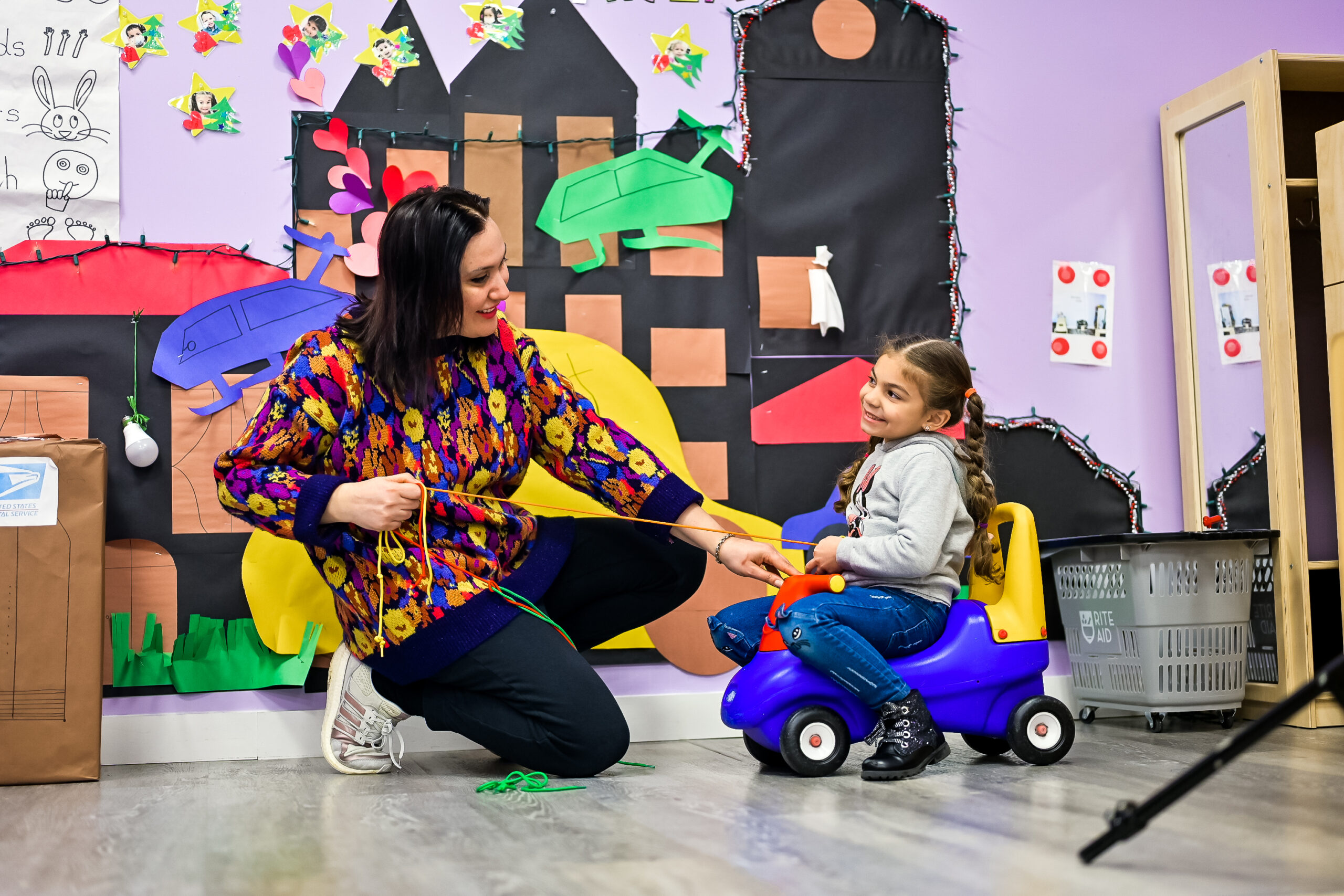 A woman with dark hair in the Little Scholars Daycare sits next to a child with a toy.