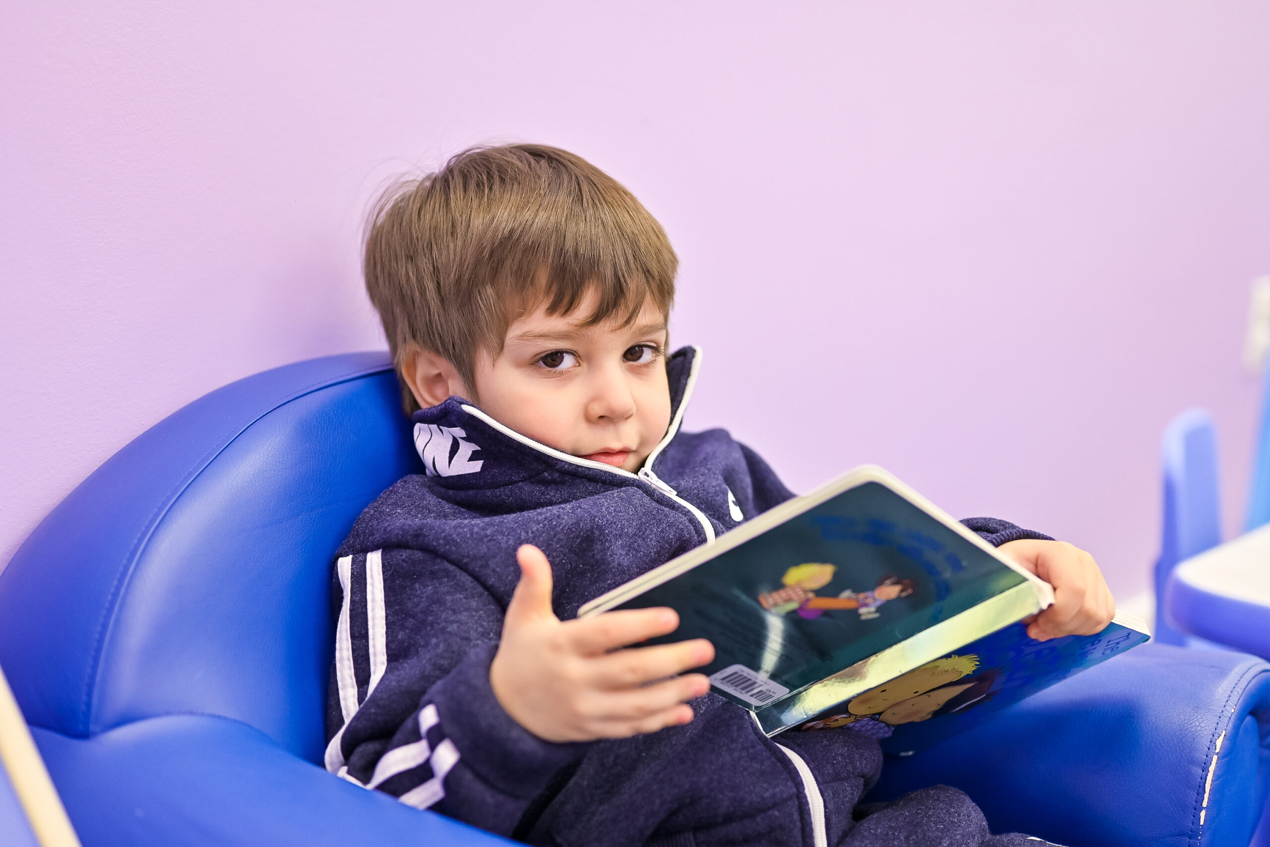 Young boy sitting in a blue chair, holding and reading a book with a focused expression in a classroom.