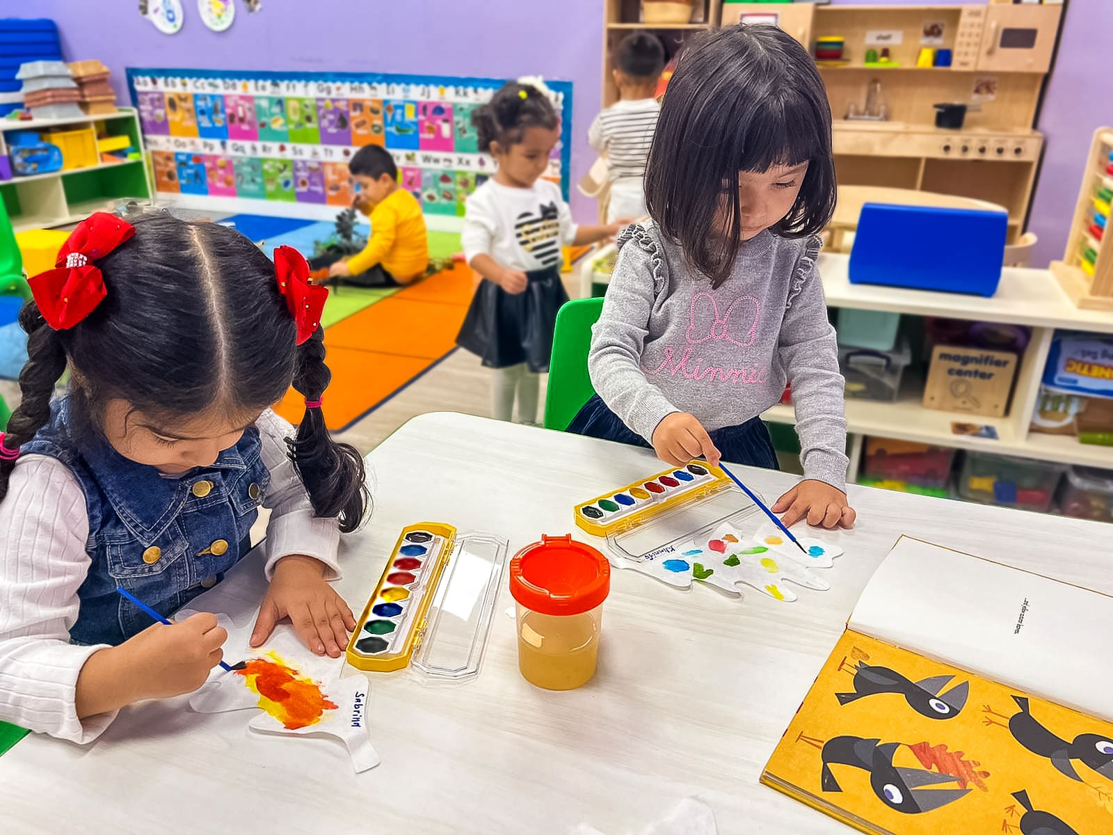 Two girls are in the garden near the table, they draw with multi-colored paints on the leaves, there are other children in the background