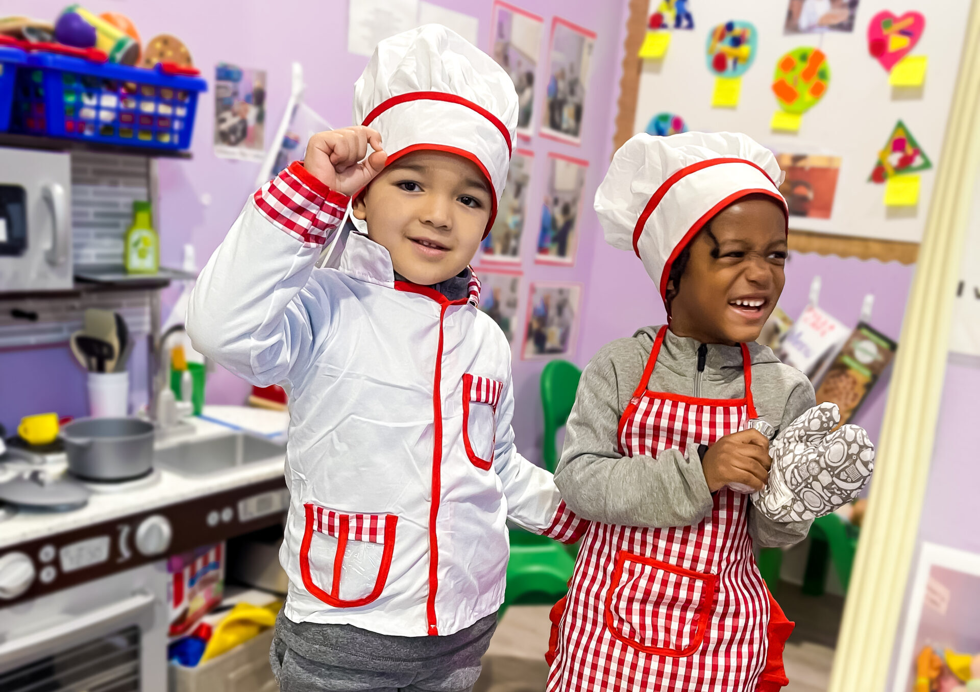 Two young children dressed as chefs, wearing white hats and aprons with red accents, smiling and posing in a play kitchen area in a daycare classroom.