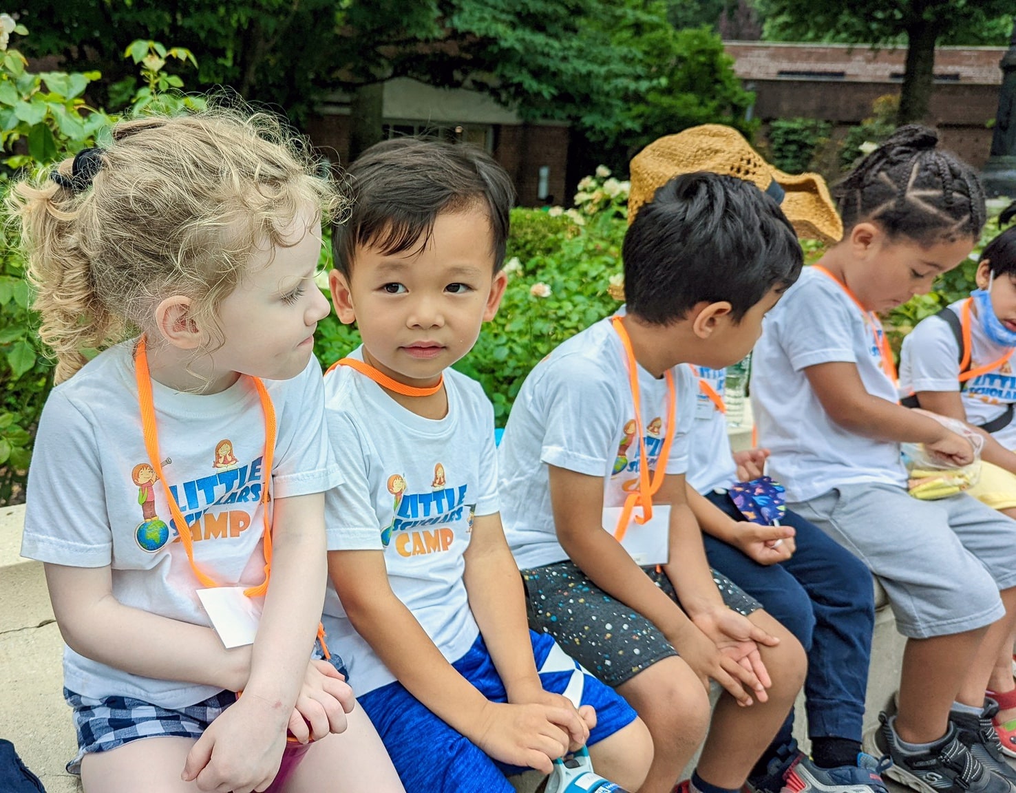 Group of preschool children with lanyards sitting together on a bench during a summer camp field trip