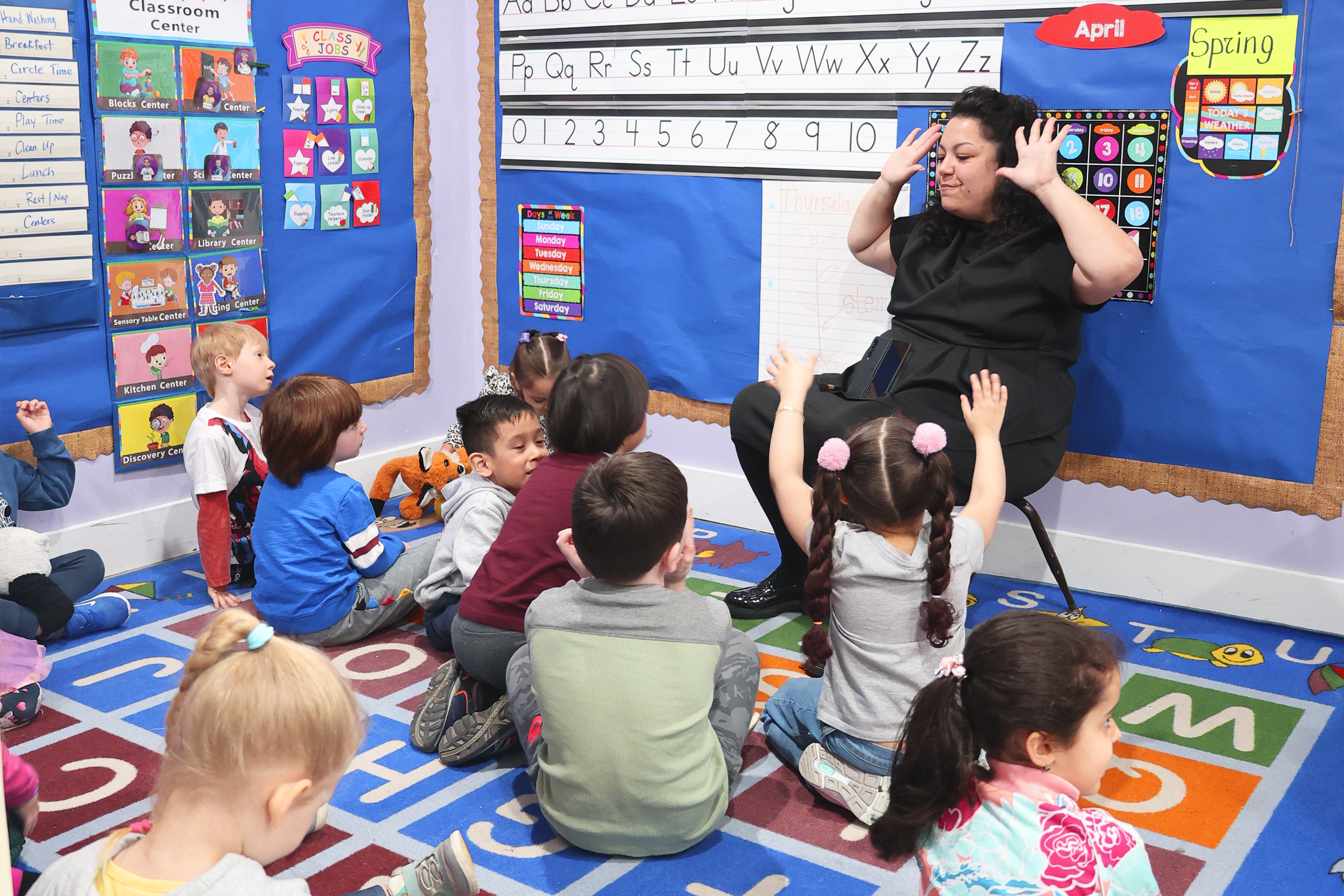 The teacher sits on a chair, around him are children in Daycare