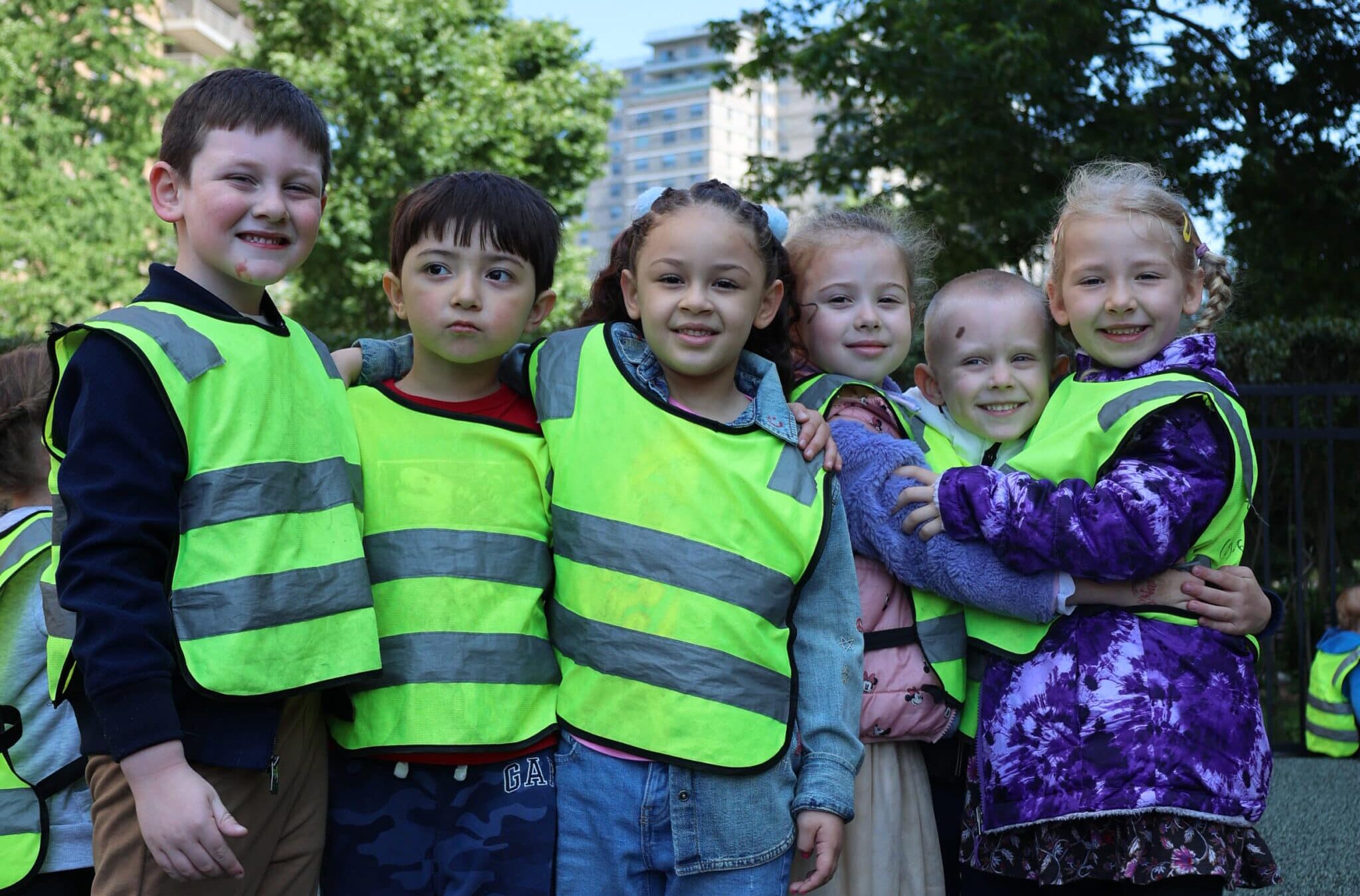 Group of five children wearing reflective safety vests, smiling and standing together outdoors in a park setting.