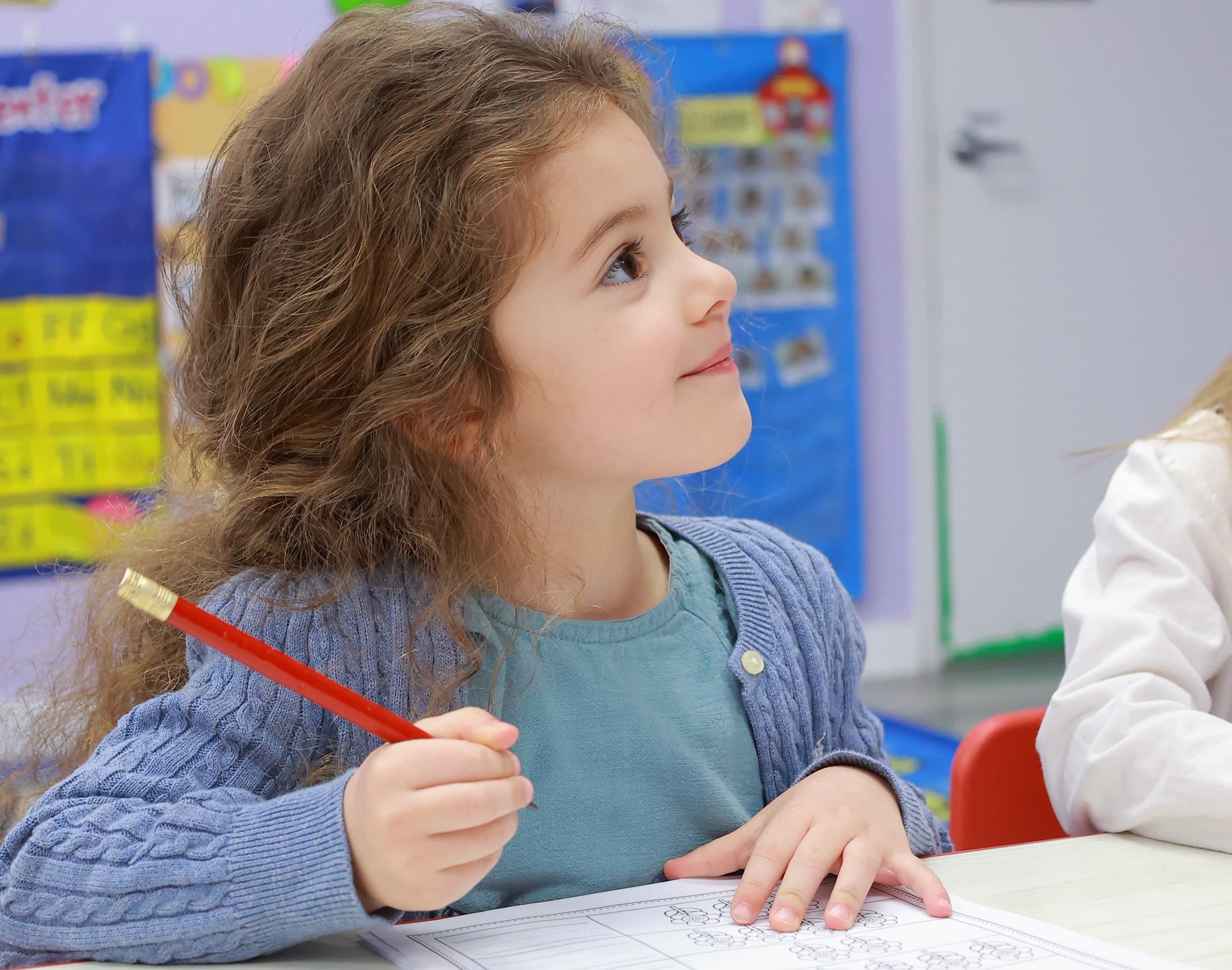 A young girl sitting at a table in a daycare classroom, holding a pencil and looking up with a thoughtful expression.