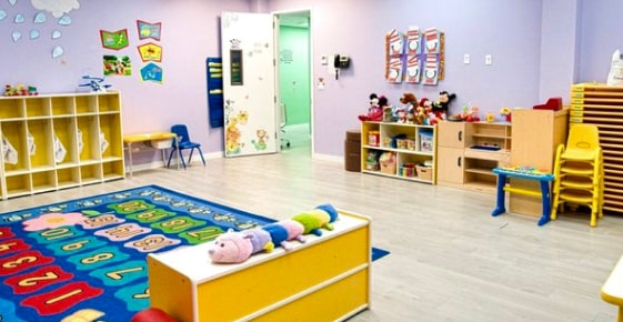 Bright and colorful daycare classroom with educational posters, shelves of toys, a number rug, and small chairs