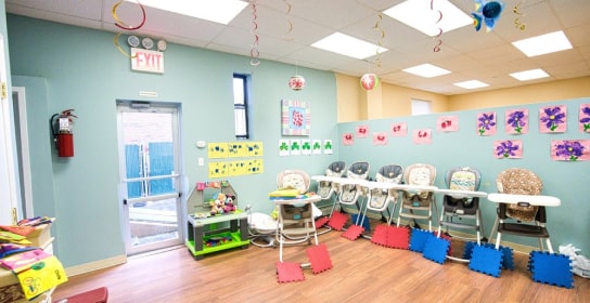 Daycare classroom with high chairs lined up against the wall, colorful artwork displayed, and foam play mats on the floor