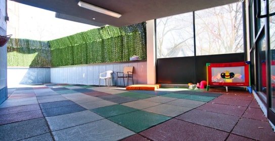 Outdoor play area at a daycare with colorful foam flooring, a playpen, and seating, enclosed by green fencing