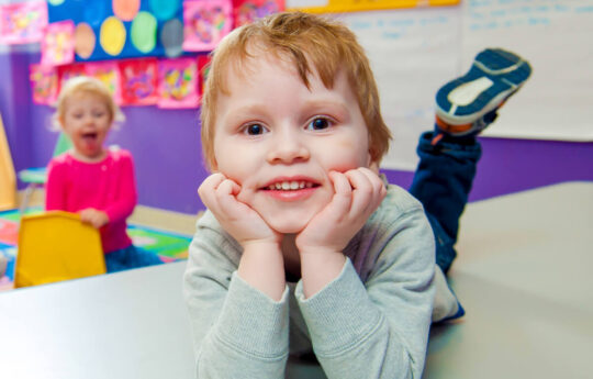 Cheerful young boy lying on a table with his chin resting on his hands in a colorful daycare classroom, with another child in the background