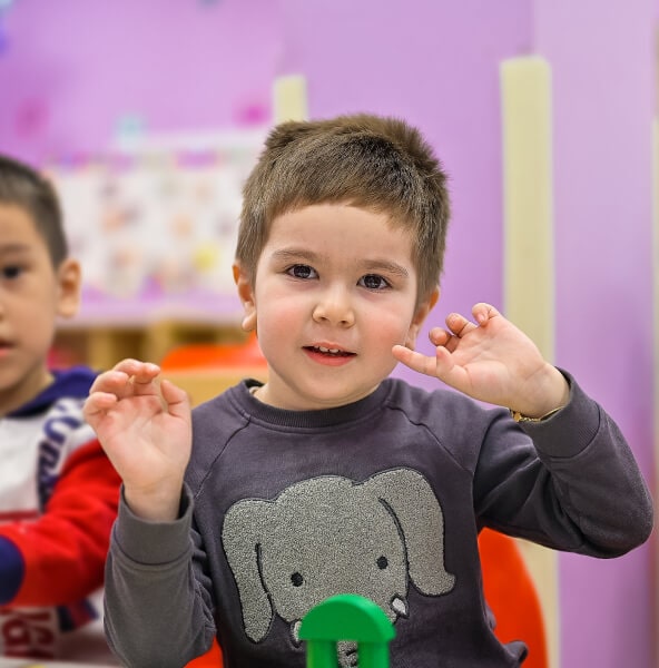 Young boy with short hair and an elephant sweater, playfully gesturing in a preschool afterschool classroom