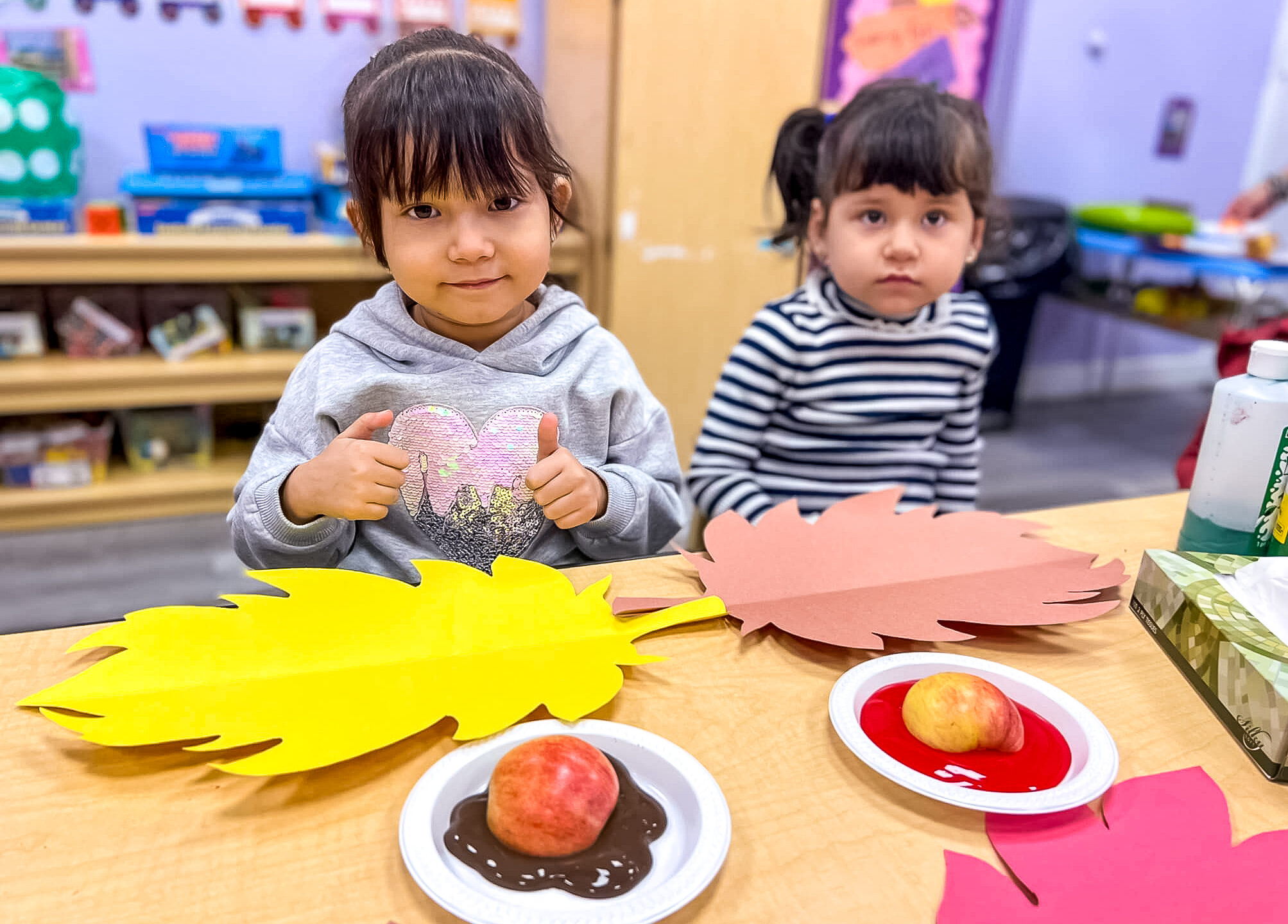 Two young girls sitting at a table in a daycare classroom, working on an apple-themed craft project.