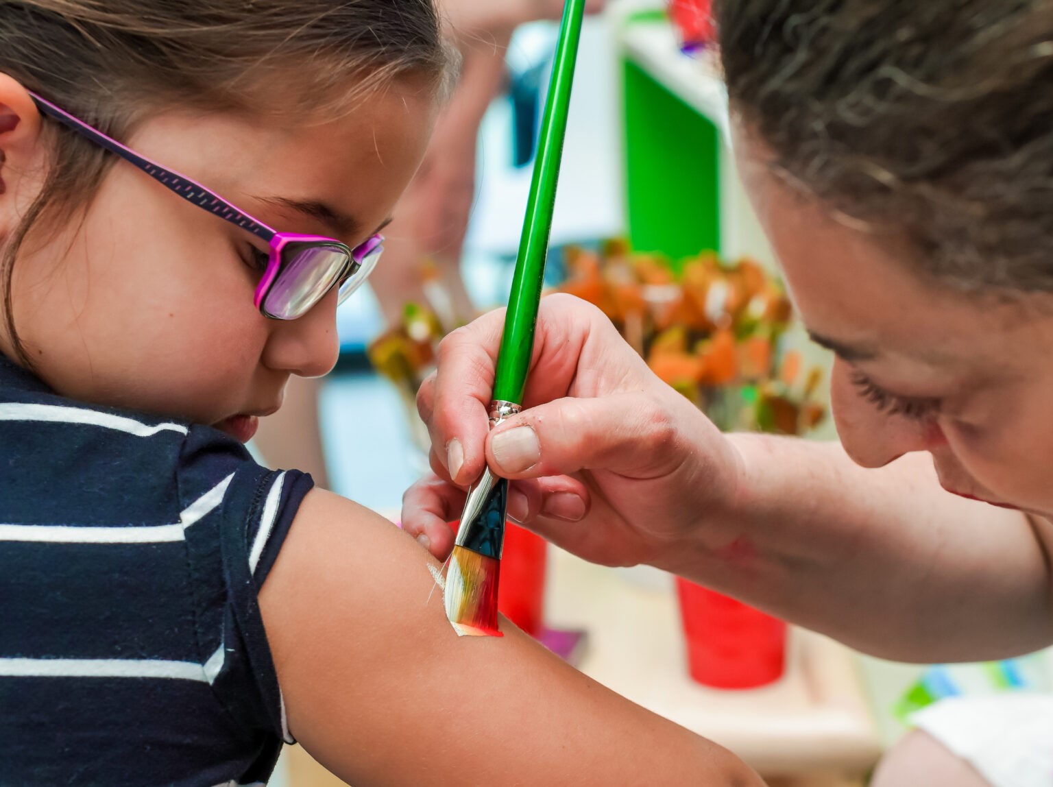 A close-up of a young girl wearing glasses getting her arm painted with a brush by an adult in a daycare classroom.