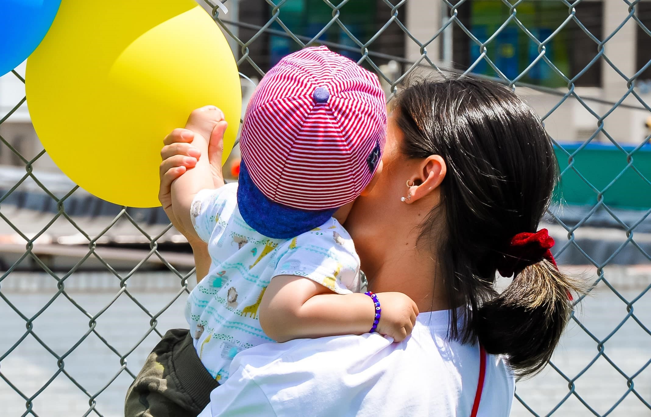 A woman holding a young child wearing a red and white striped hat, with the child reaching towards a yellow balloon tied to a fence.