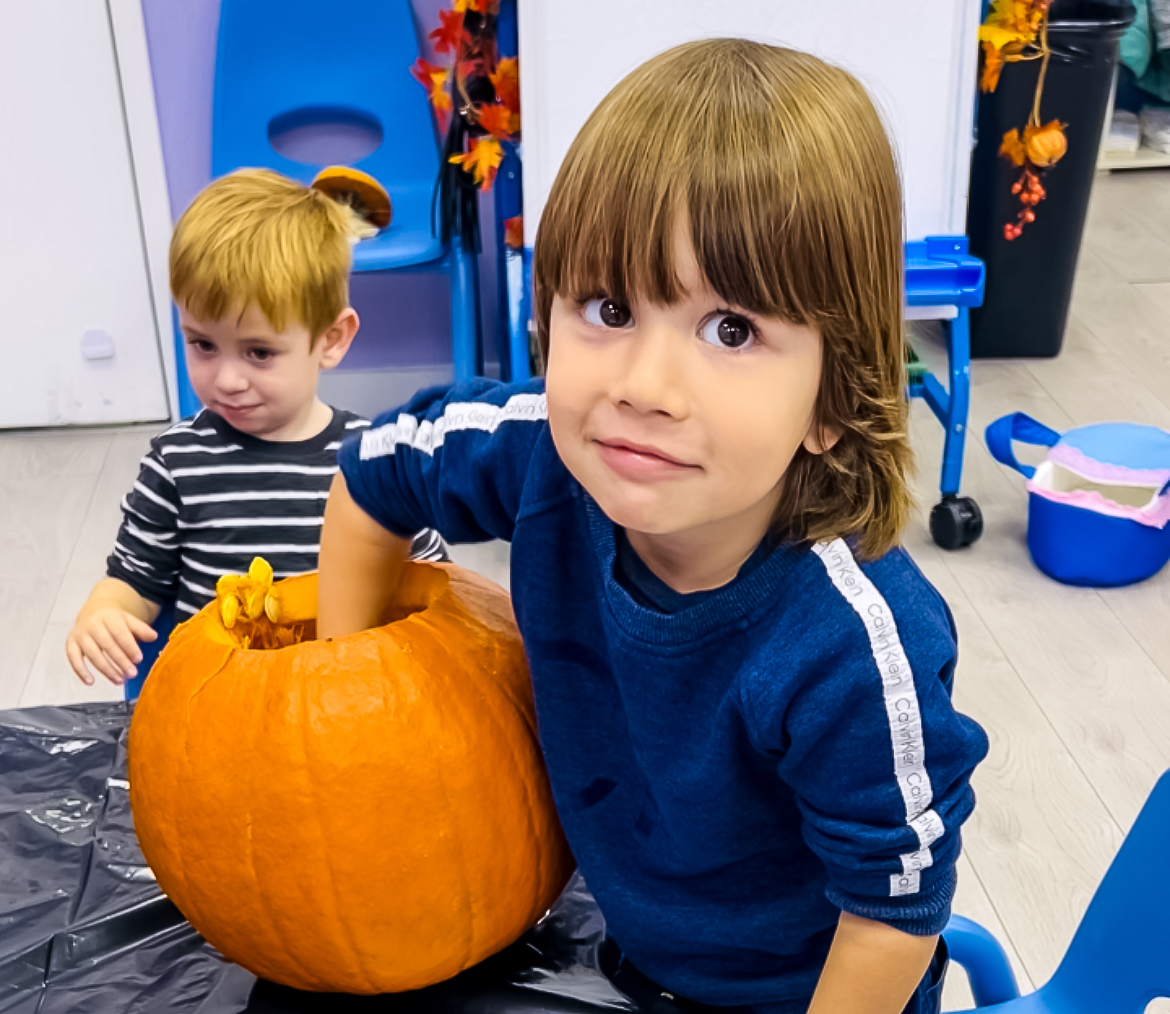 Two young boys in a classroom, one with his hand inside a large pumpkin, preparing for Halloween activities.