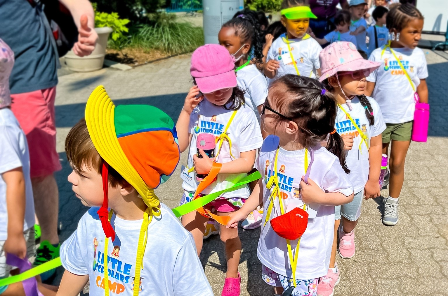 A group of young children wearing matching white T-shirts and colorful hats, walking in a line while holding a green rope during a field trip.