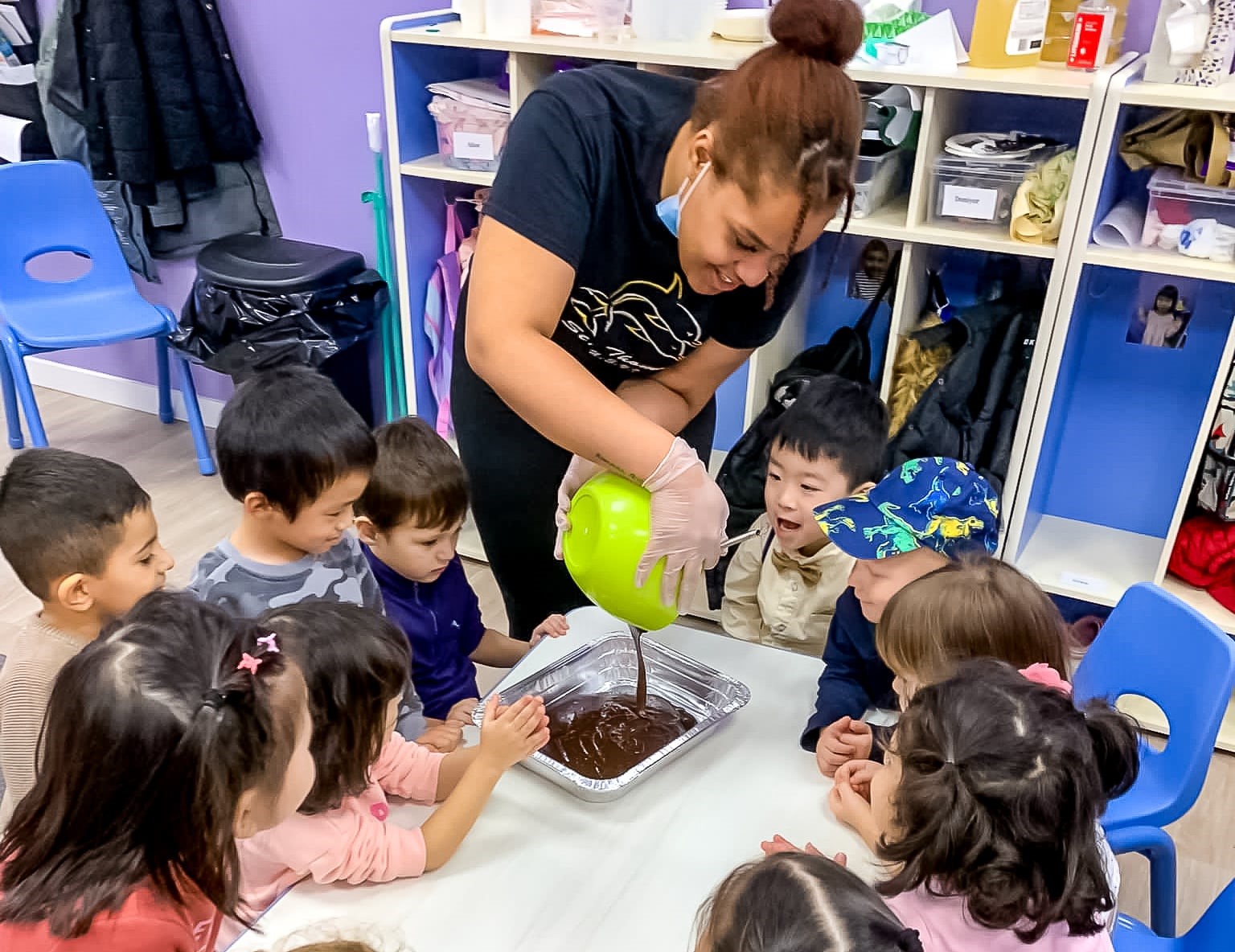 Group of children gathered around a table, watching a teacher pour a mixture into a tray during a cooking activity in a classroom