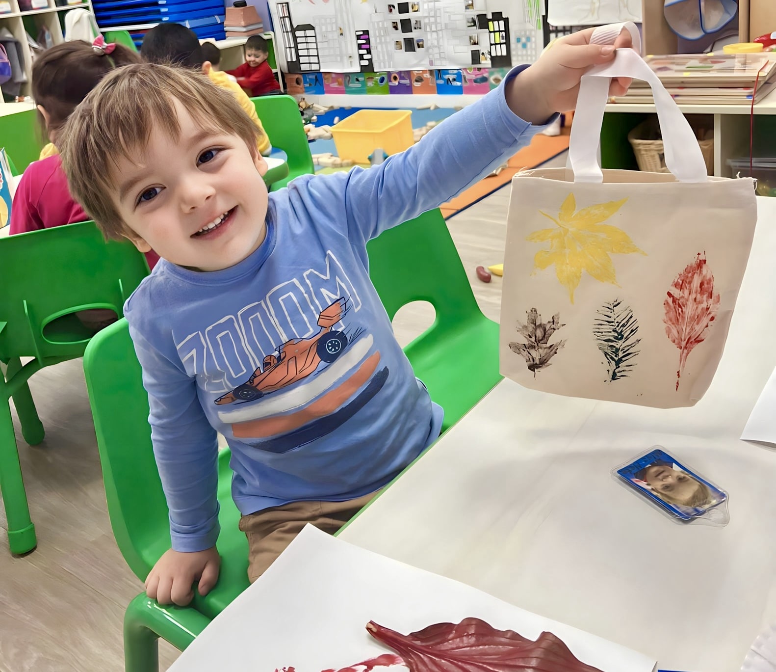 A young boy sitting at a table in a daycare classroom, proudly holding up a decorated canvas bag with leaf prints.