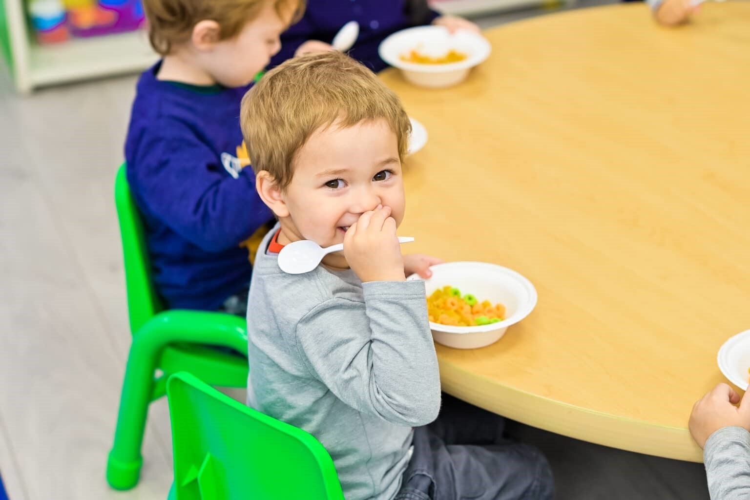 A young boy sitting at a round table in a daycare classroom, smiling and eating from a bowl with a spoon.