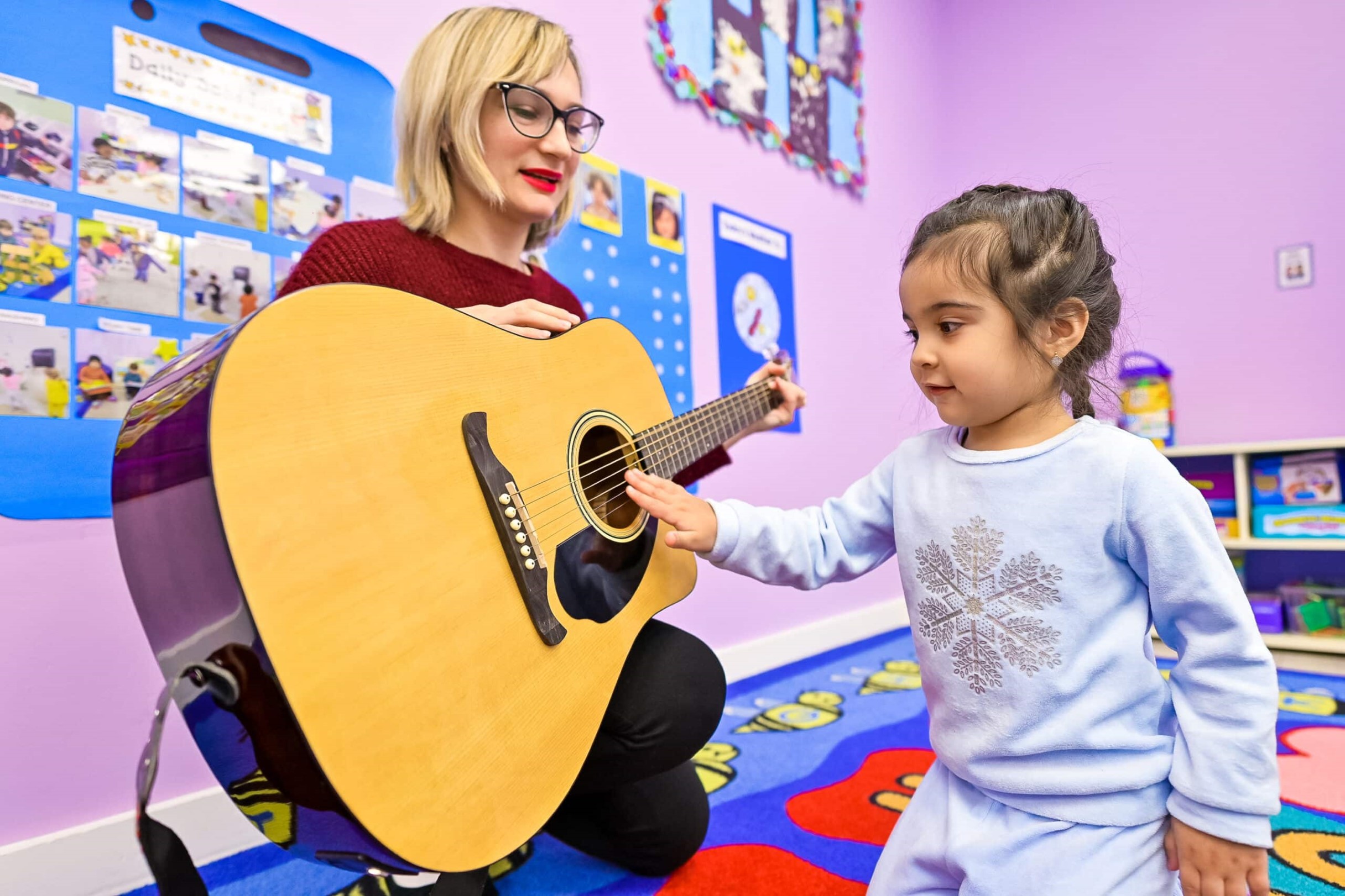 Young girl in a light blue sweater touching the strings of a guitar played by a teacher with blonde hair and glasses, in a colorful classroom setting