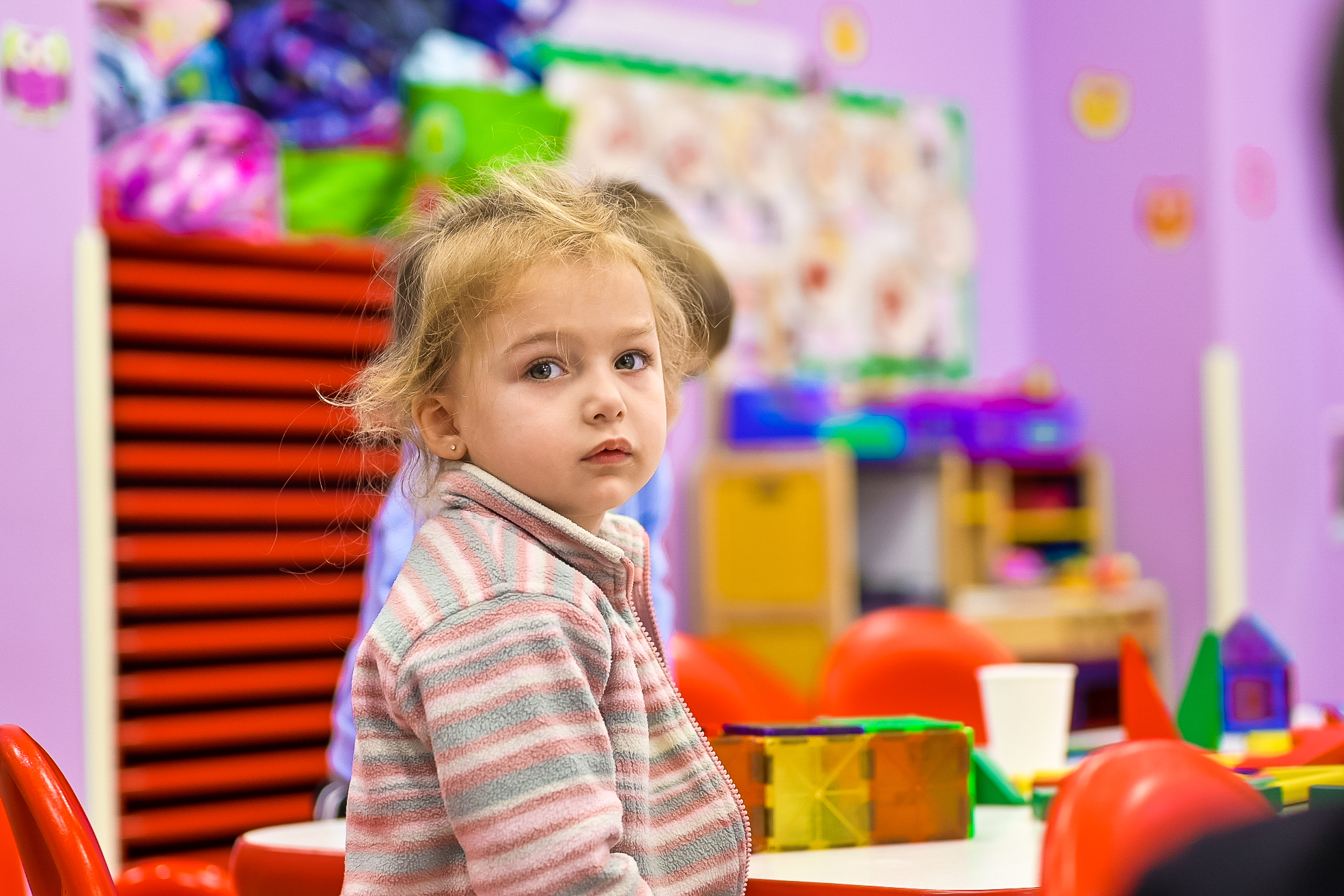 A young girl wearing a striped sweater, sitting at a table in a daycare classroom, looking off to the side with a thoughtful expression.