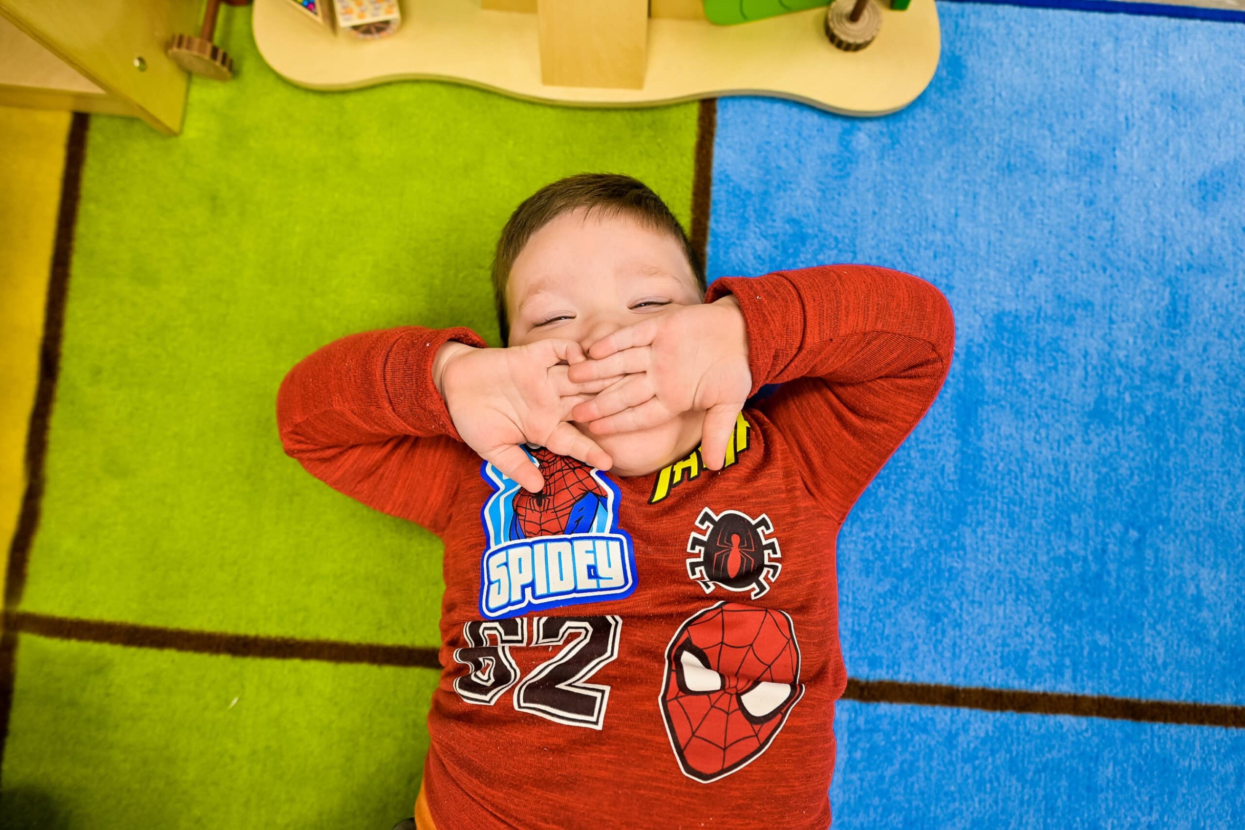 A young boy lying on a colorful rug in a daycare classroom, wearing a red shirt with Spider-Man graphics.