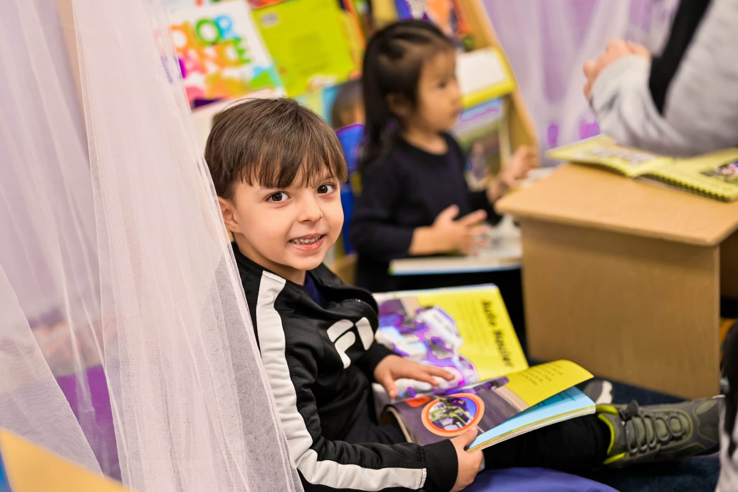 Young boy sitting in a reading corner with a book in his lap, smiling at the camera, while another child reads in the background in a colorful classroom