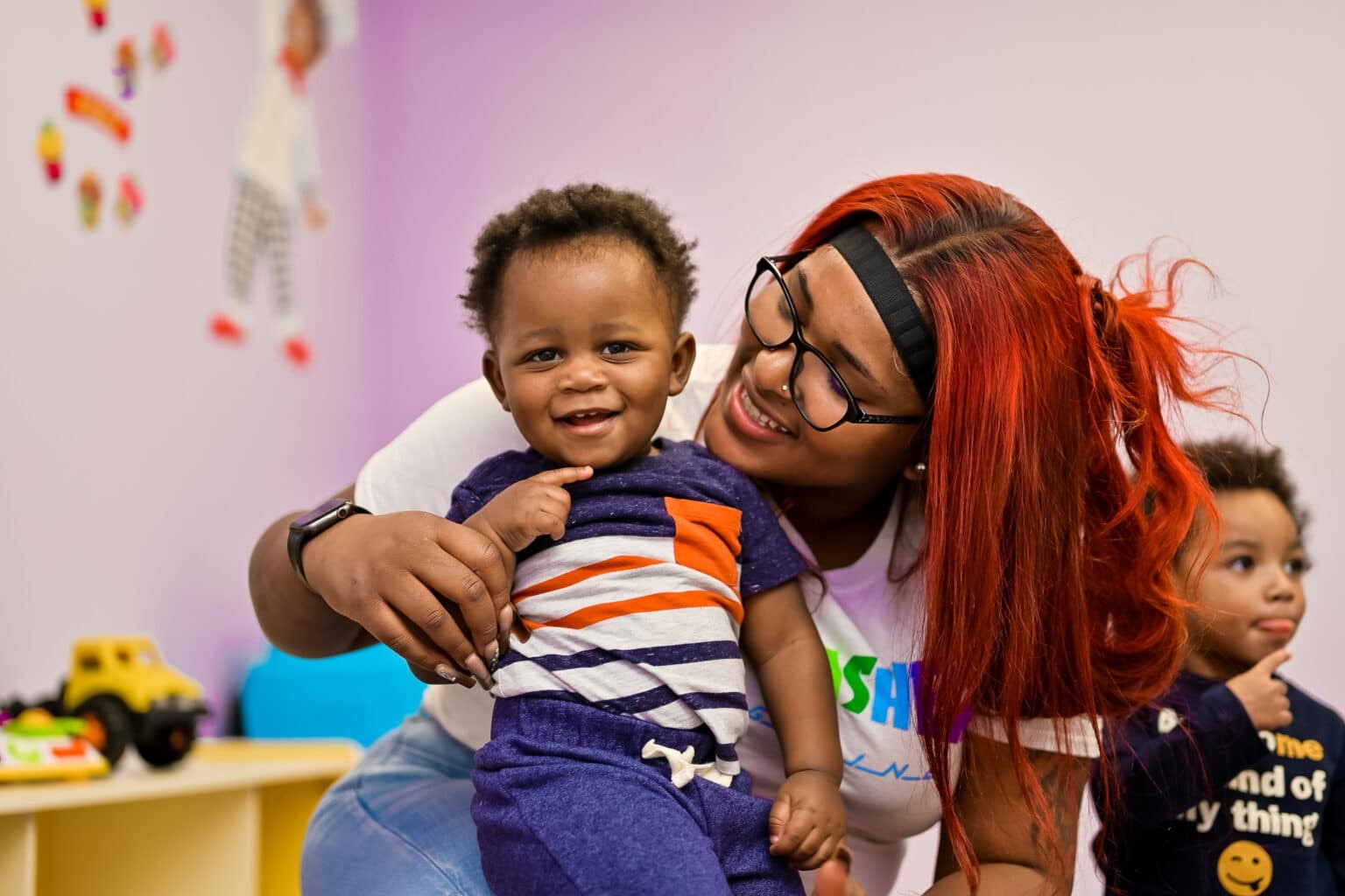 Smiling teacher with red hair and glasses holding a happy toddler in a classroom, with another child in the background