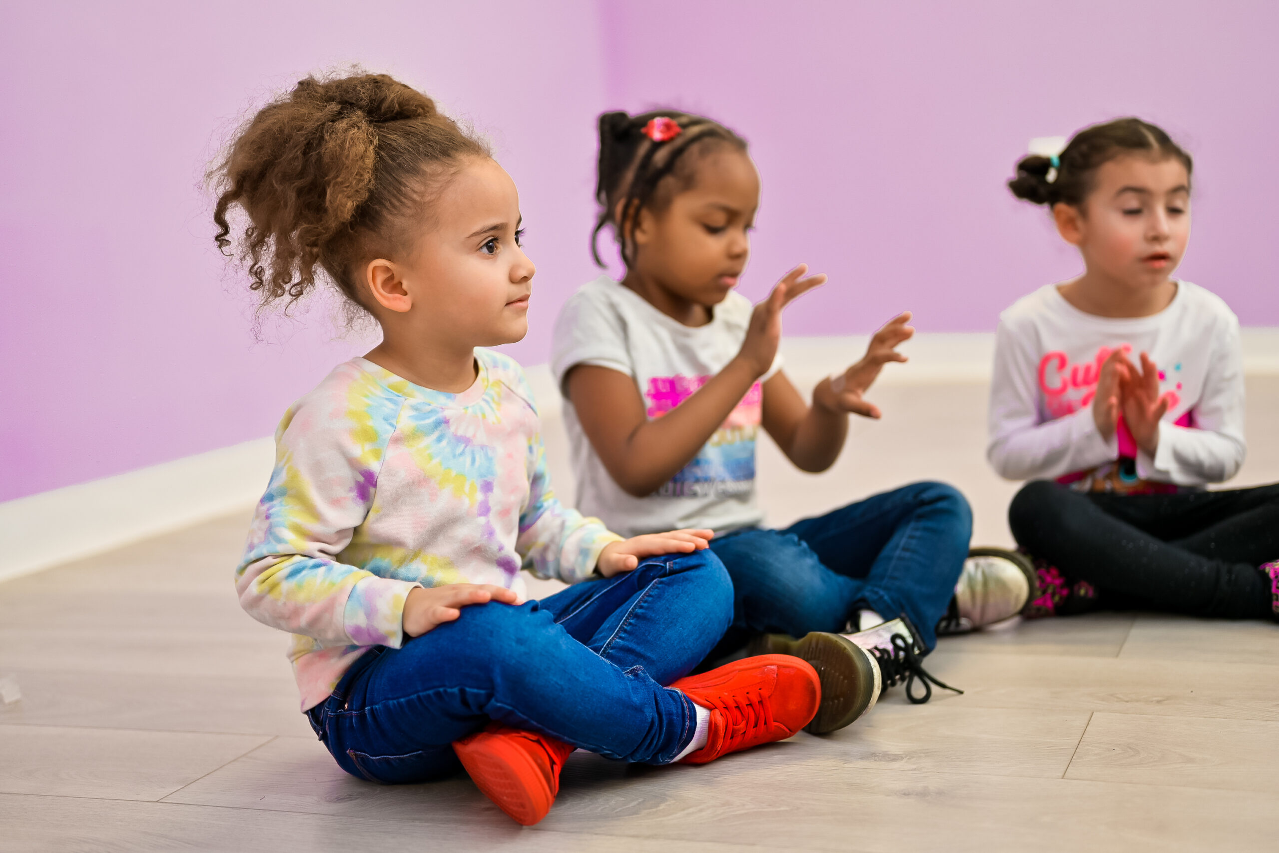 Three young girls sitting cross-legged on the floor of a daycare classroom, engaged in an activity together.