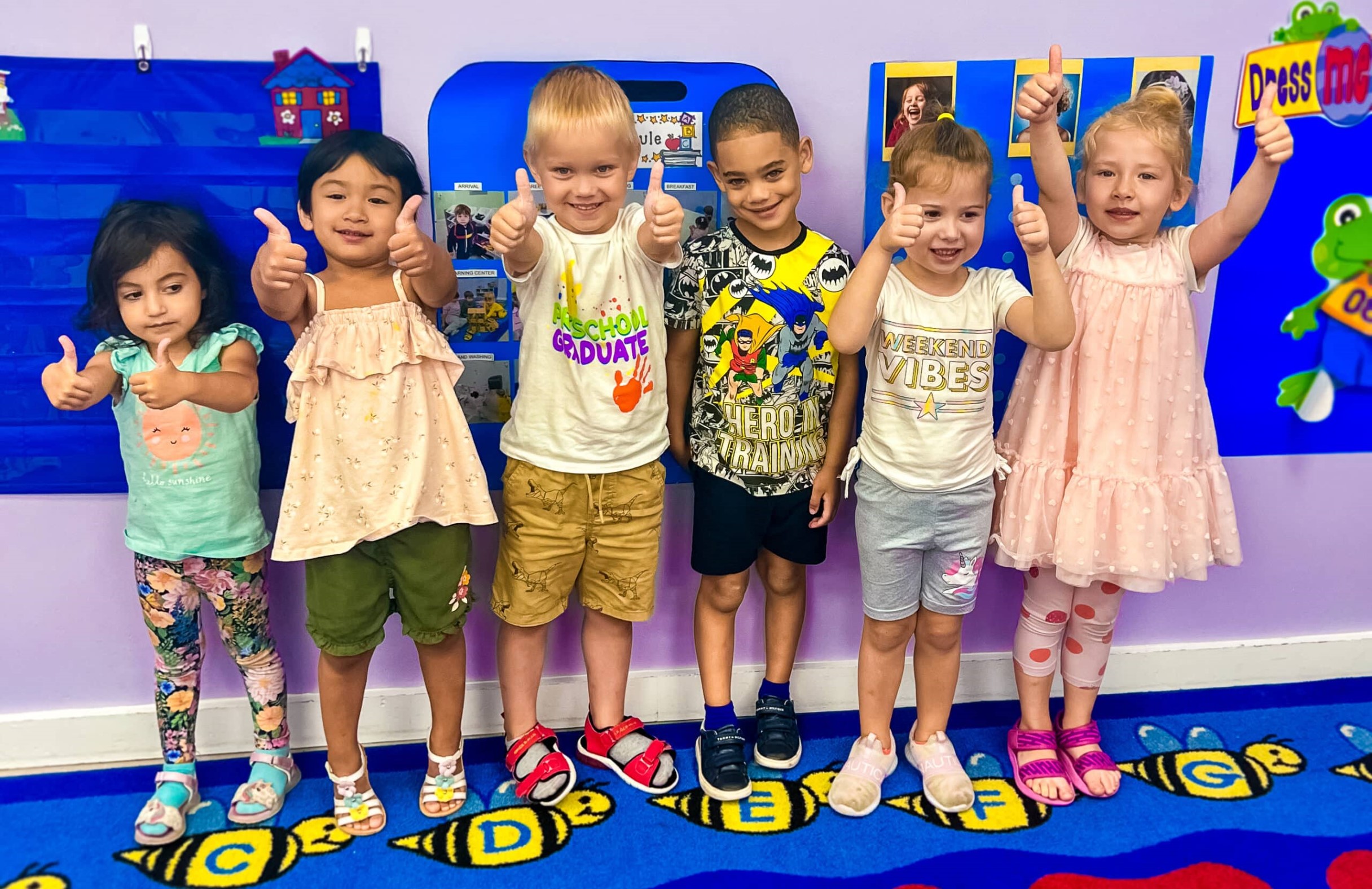 A group of six young children standing in a row on a colorful alphabet rug in a daycare classroom, smiling and giving thumbs up.