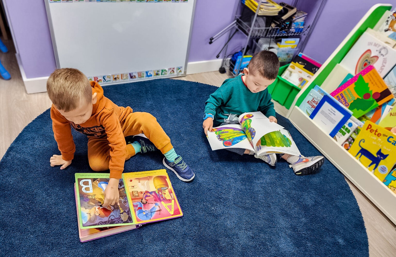 Two young boys sitting on a blue carpet in a classroom, each reading a book from the classroom's bookshelf