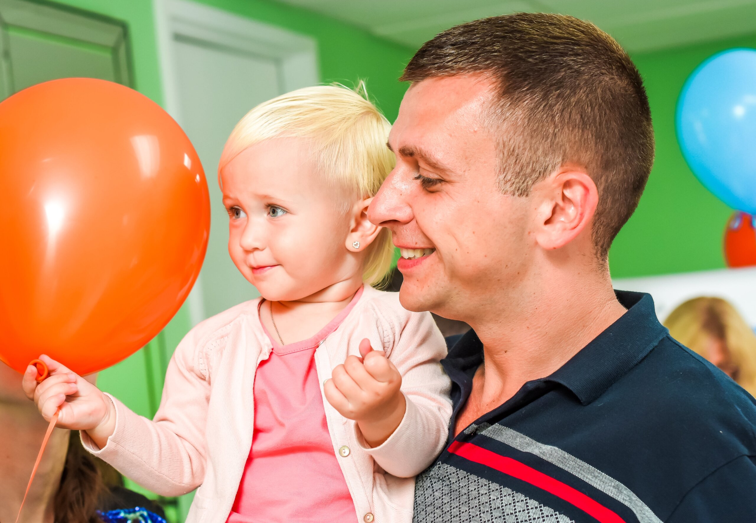 Smiling parent holding a young child with blonde hair and an orange balloon, both looking happy in a brightly colored room