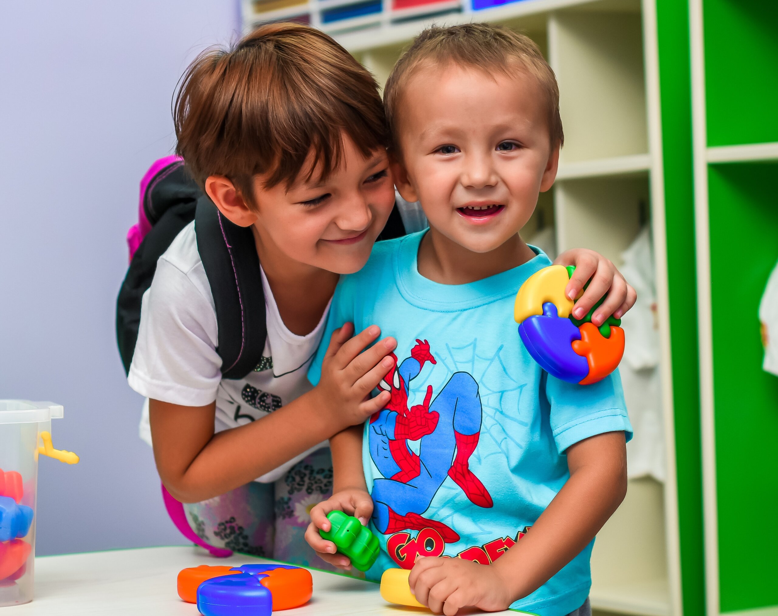 Two smiling children playing with colorful toys at a table in a classroom, with shelves and storage bins in the background