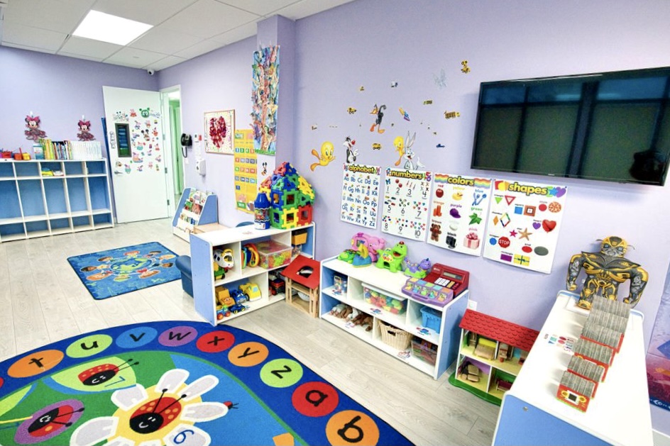 Colorful daycare classroom with educational posters, toy shelves, a TV, and an alphabet rug on the floor.