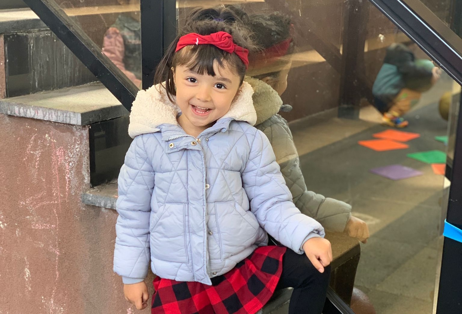 Young girl in a gray jacket and red plaid skirt, smiling while standing outside the daycare with colorful paper shapes on the floor in the background.