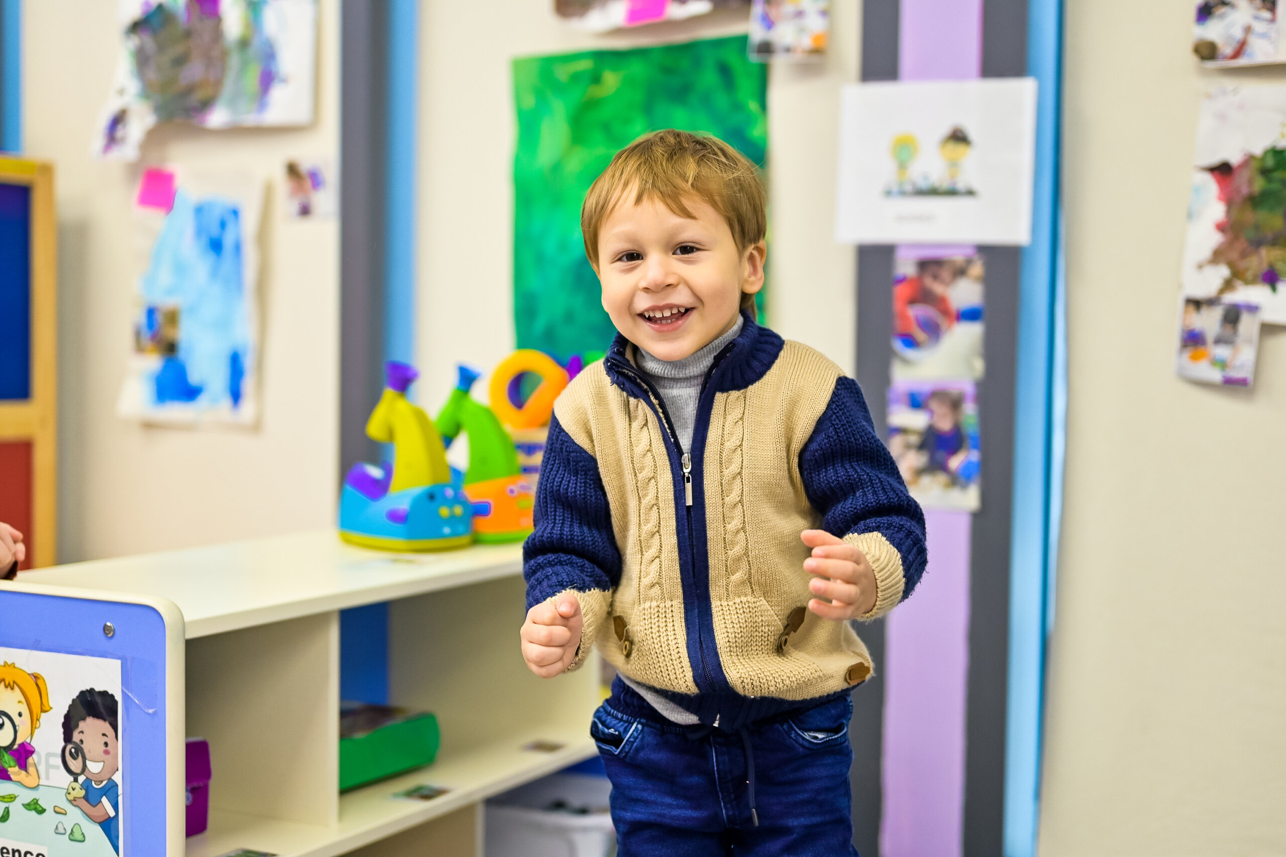 A young boy smiling and standing in a daycare classroom, wearing a beige and navy blue sweater.