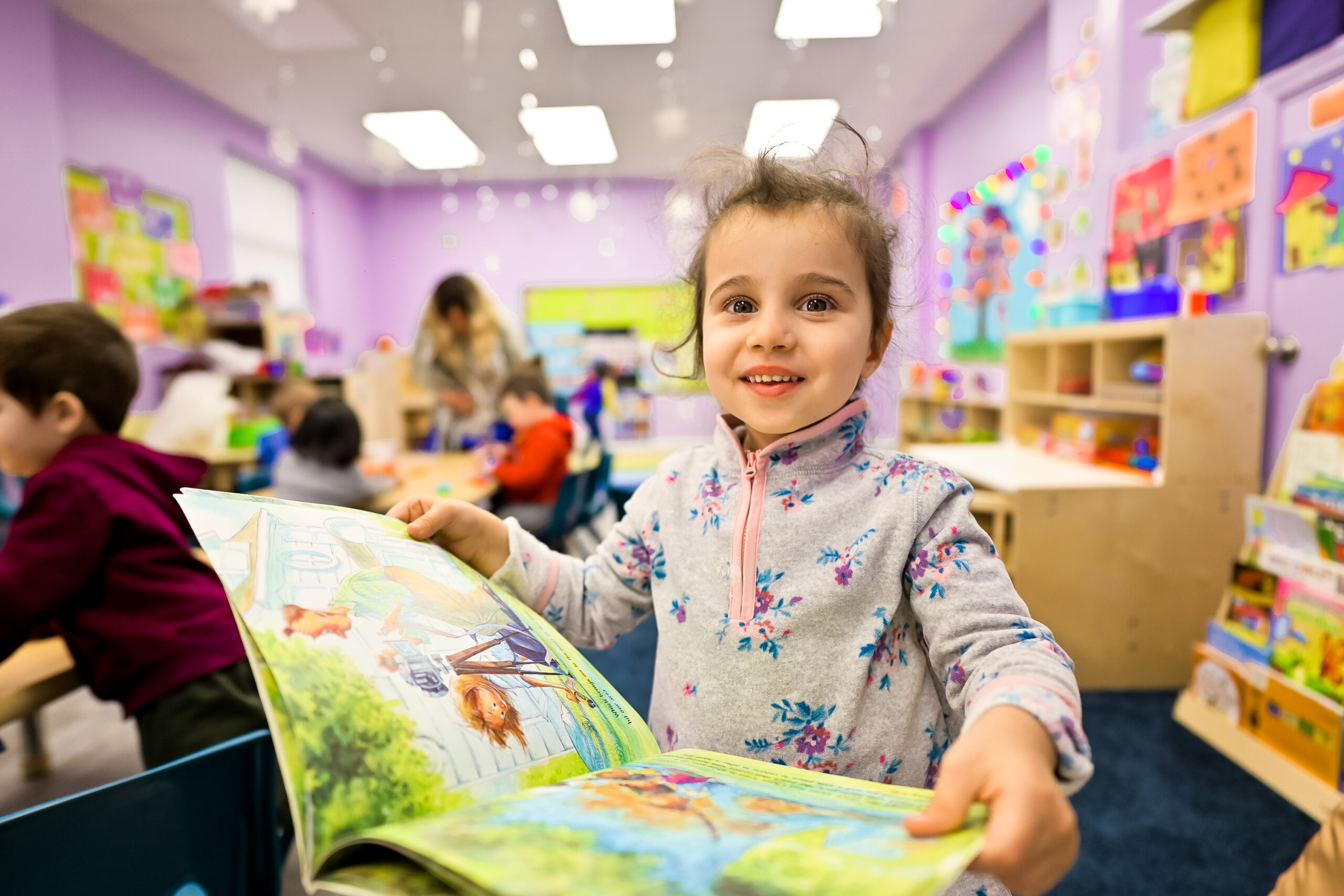 Young girl smiling and holding an open picture book in a colorful classroom with other children and educational materials in the background.