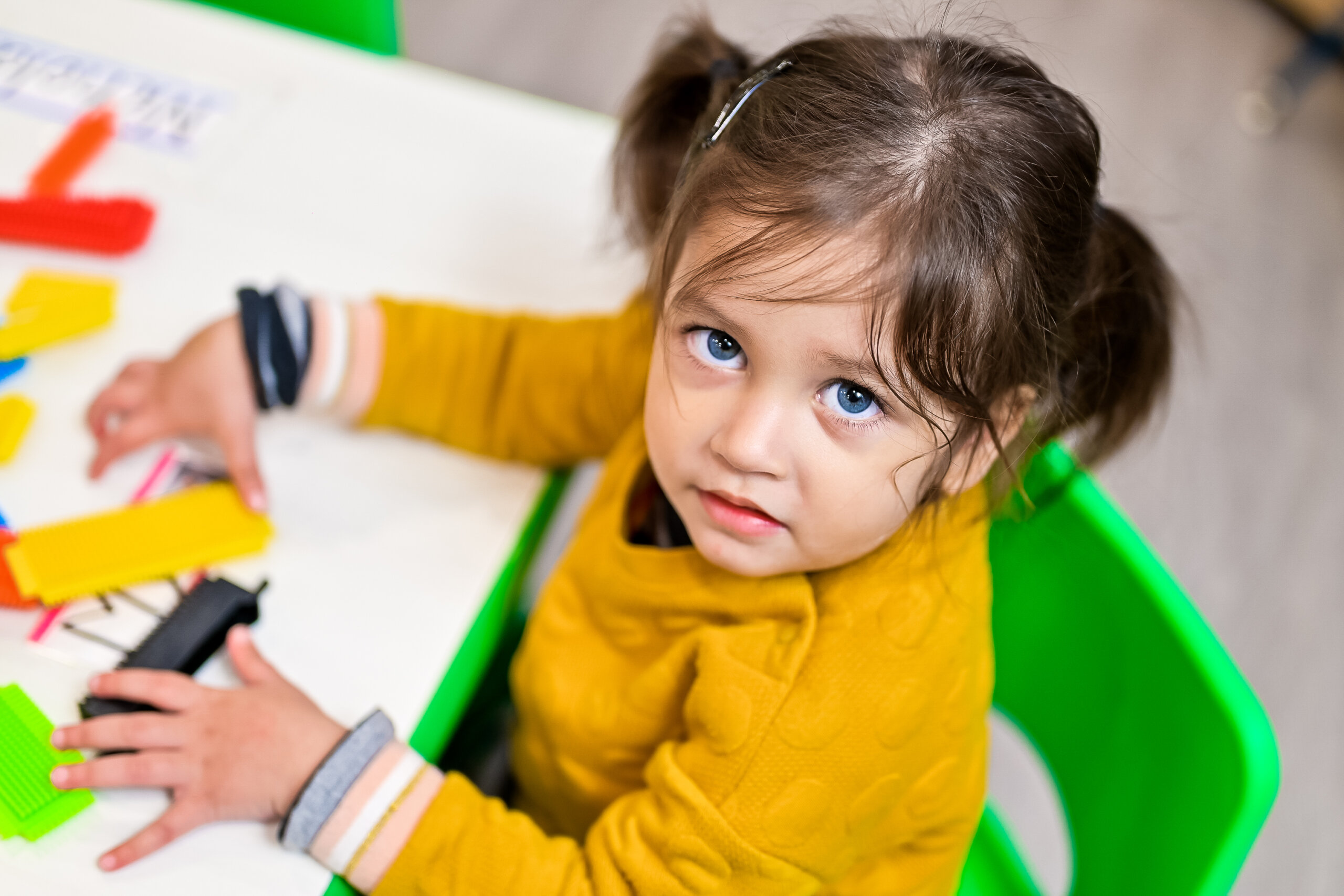 A young girl with pigtails sitting at a table in a daycare classroom, looking up at the camera while playing with colorful construction toys.