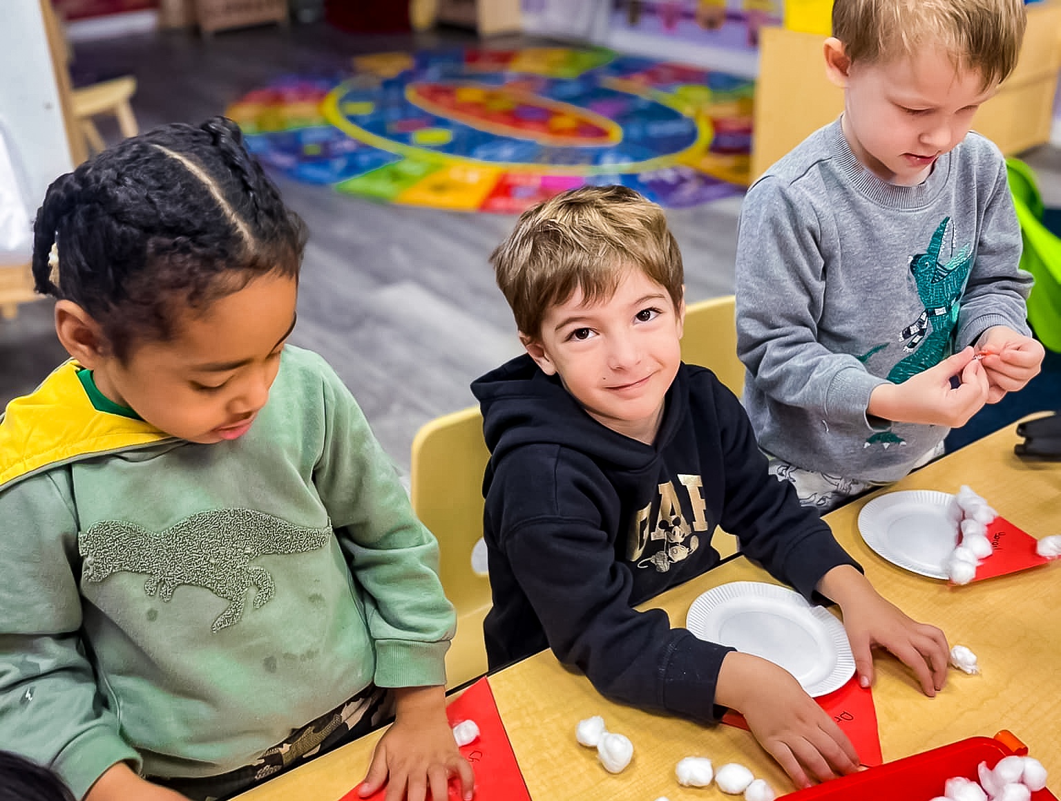 Three young children sitting at a table, engaged in a crafting activity with cotton balls and paper plates in a classroom.