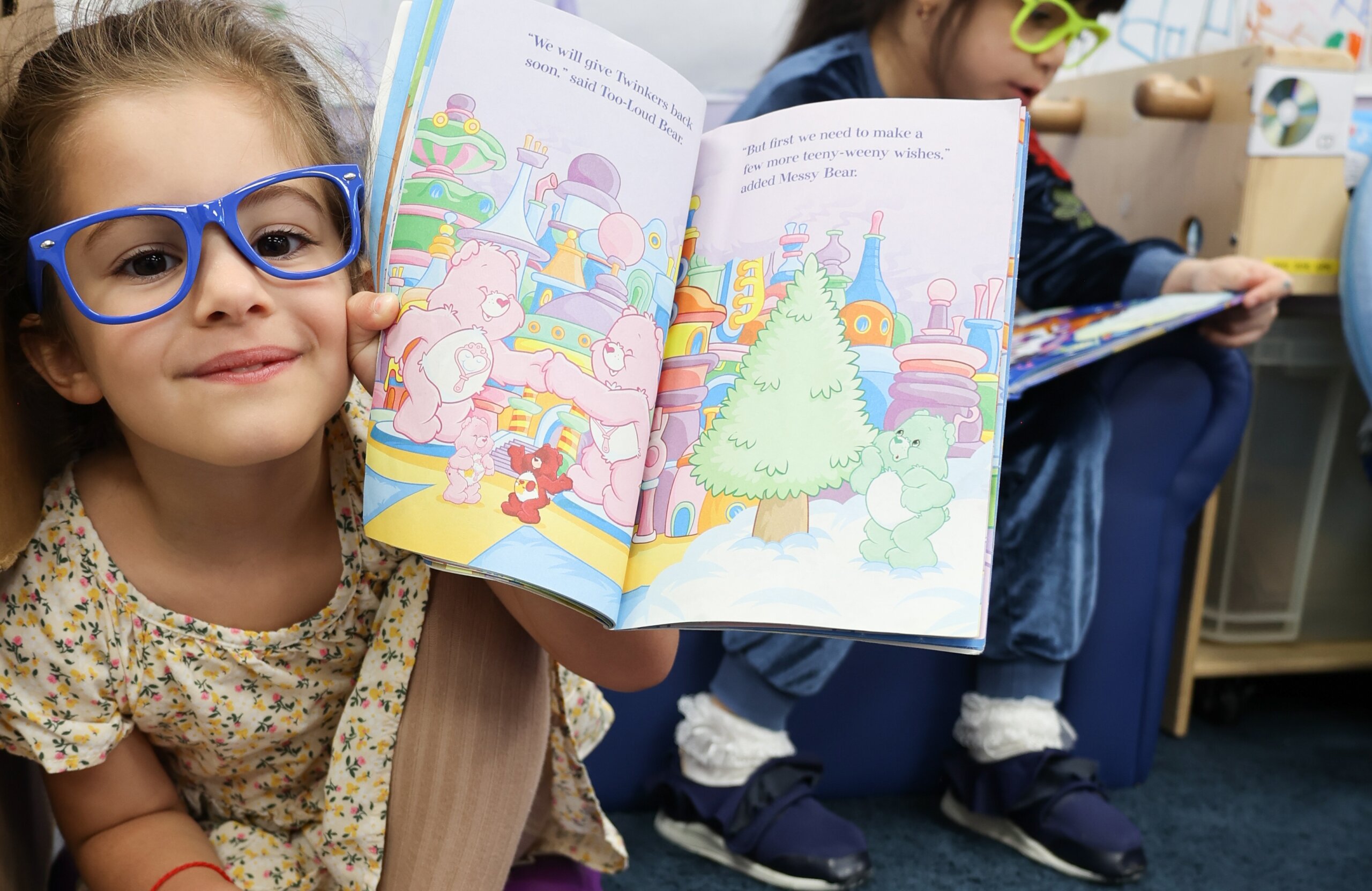 Young girl wearing blue glasses, smiling and holding up an open picture book with colorful illustrations, while another child reads in the background.