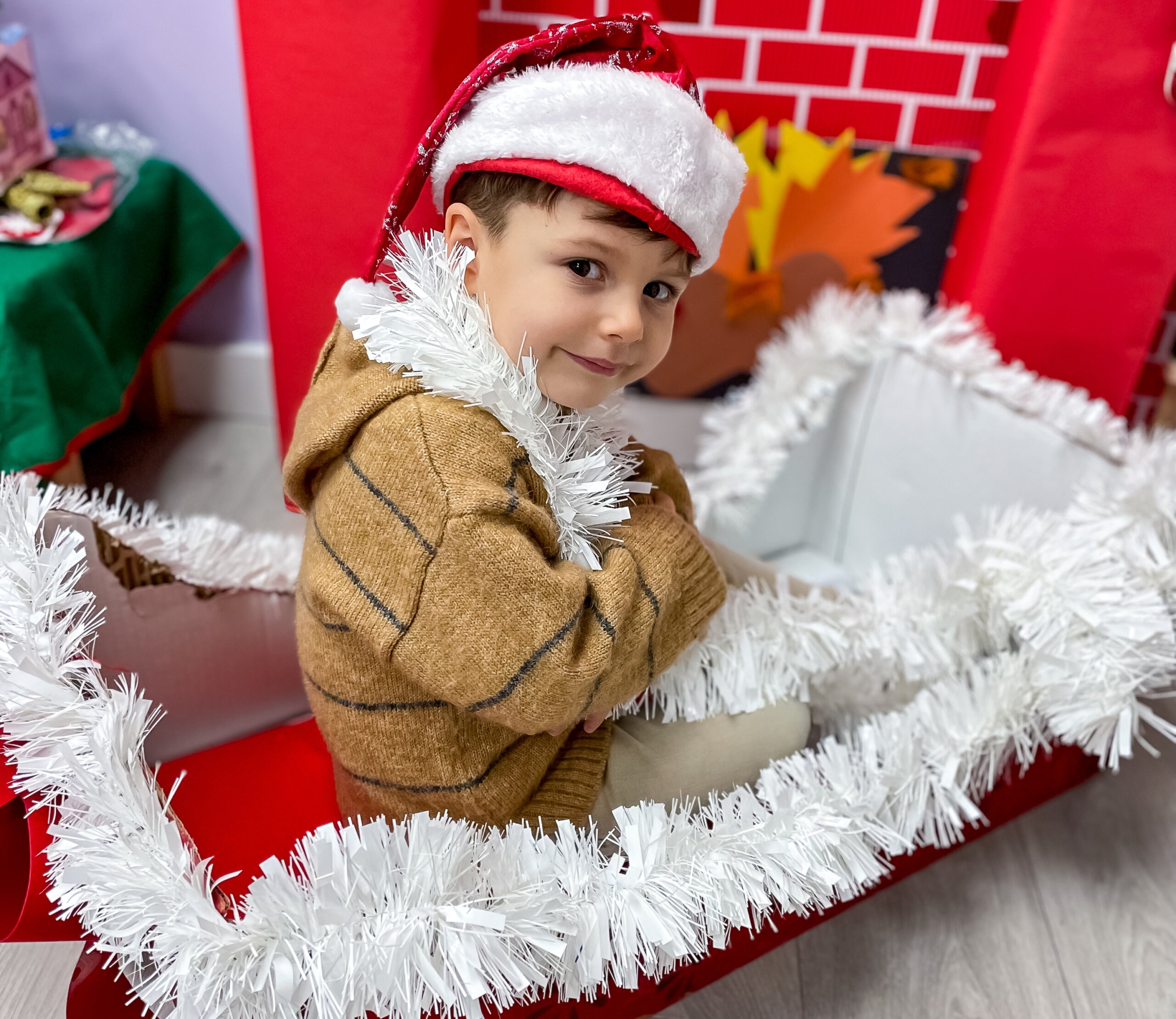 Young boy wearing a Santa hat and a brown sweater, surrounded by white tinsel, sitting in a festive holiday setting with a red background and fireplace decoration