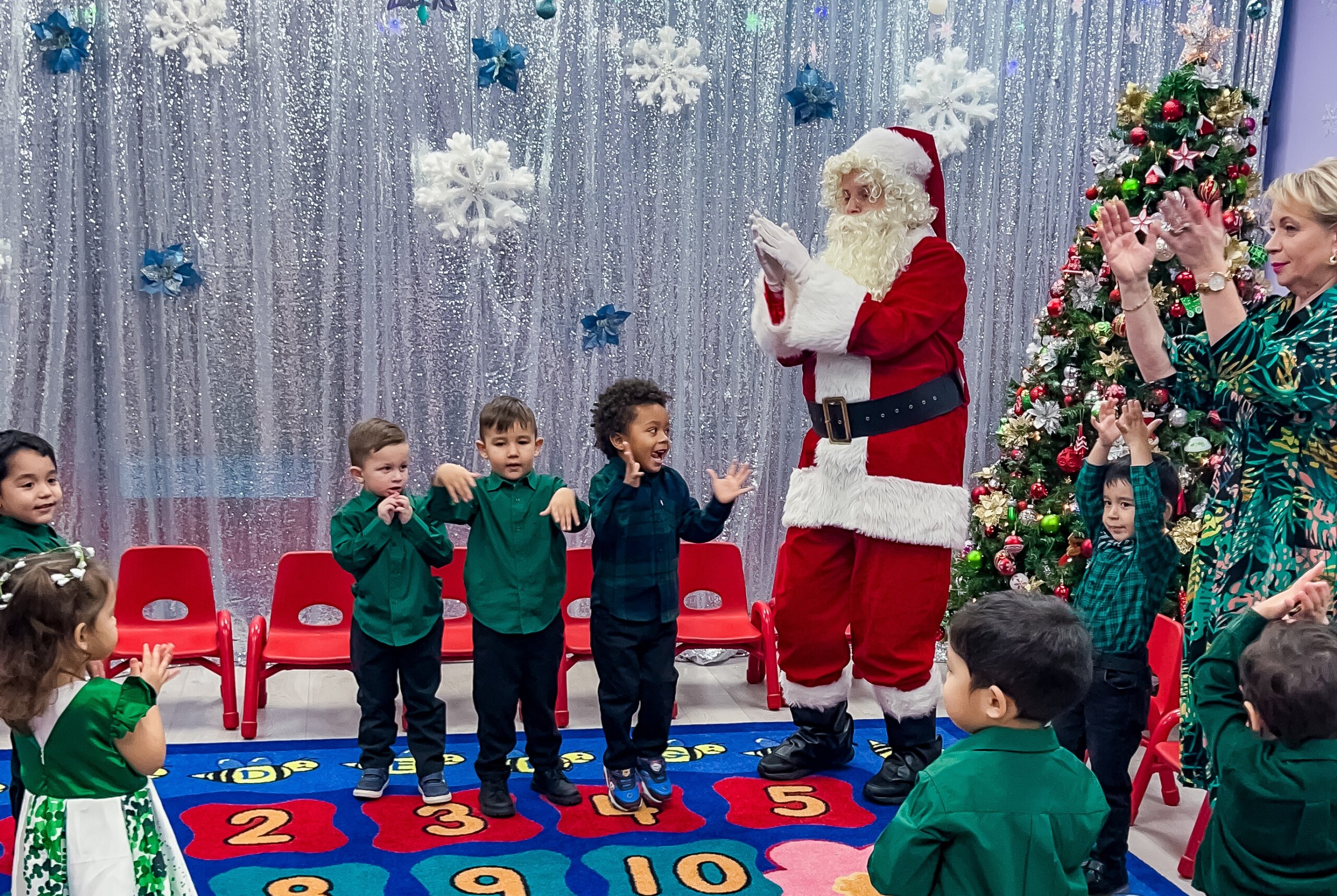 Group of young children dressed in green shirts, standing in a classroom with holiday decorations, interacting with a person dressed as Santa Claus and a teacher