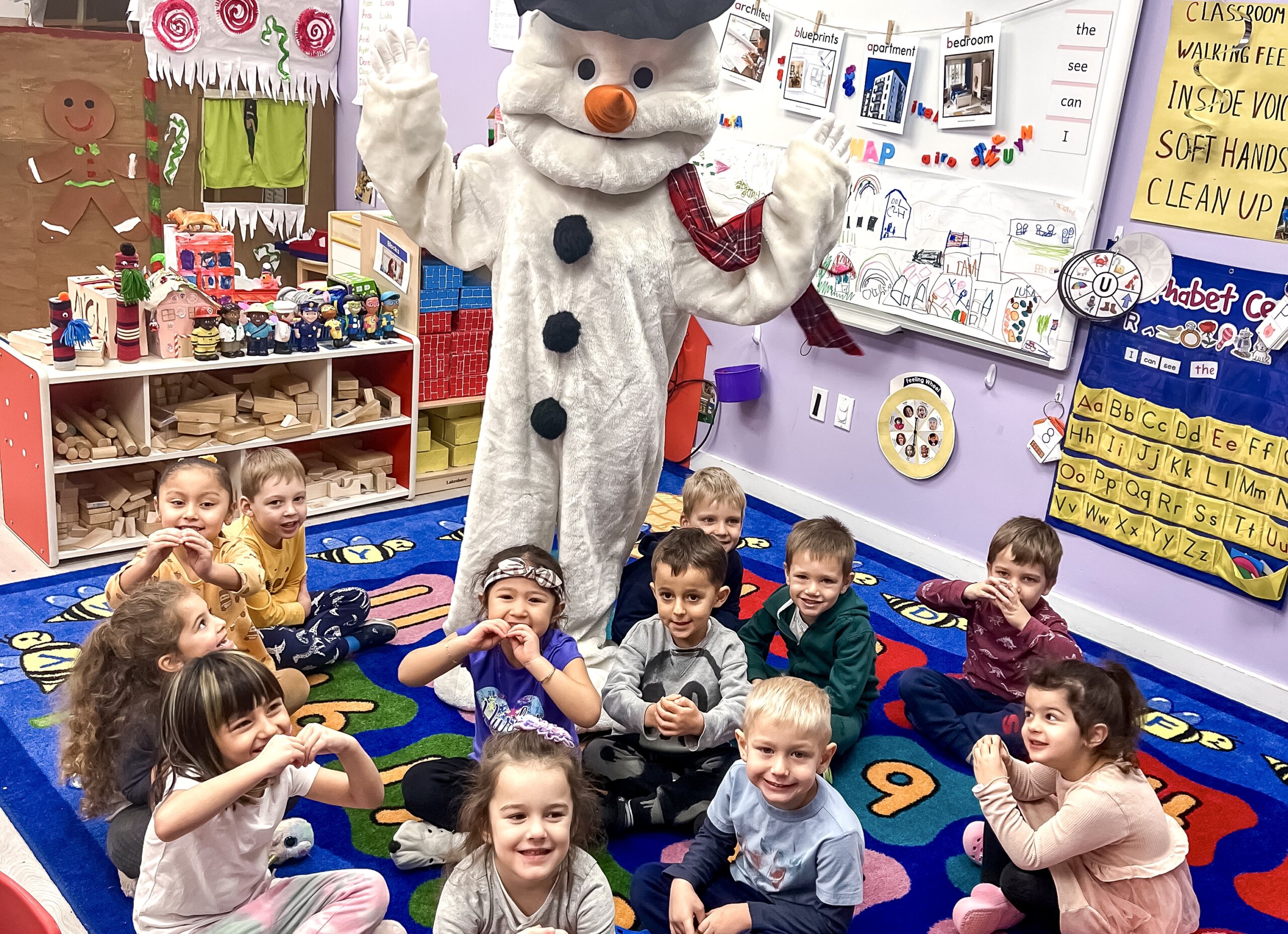 Group of children sitting on a colorful rug in a daycare classroom, posing with a person dressed in a snowman costume