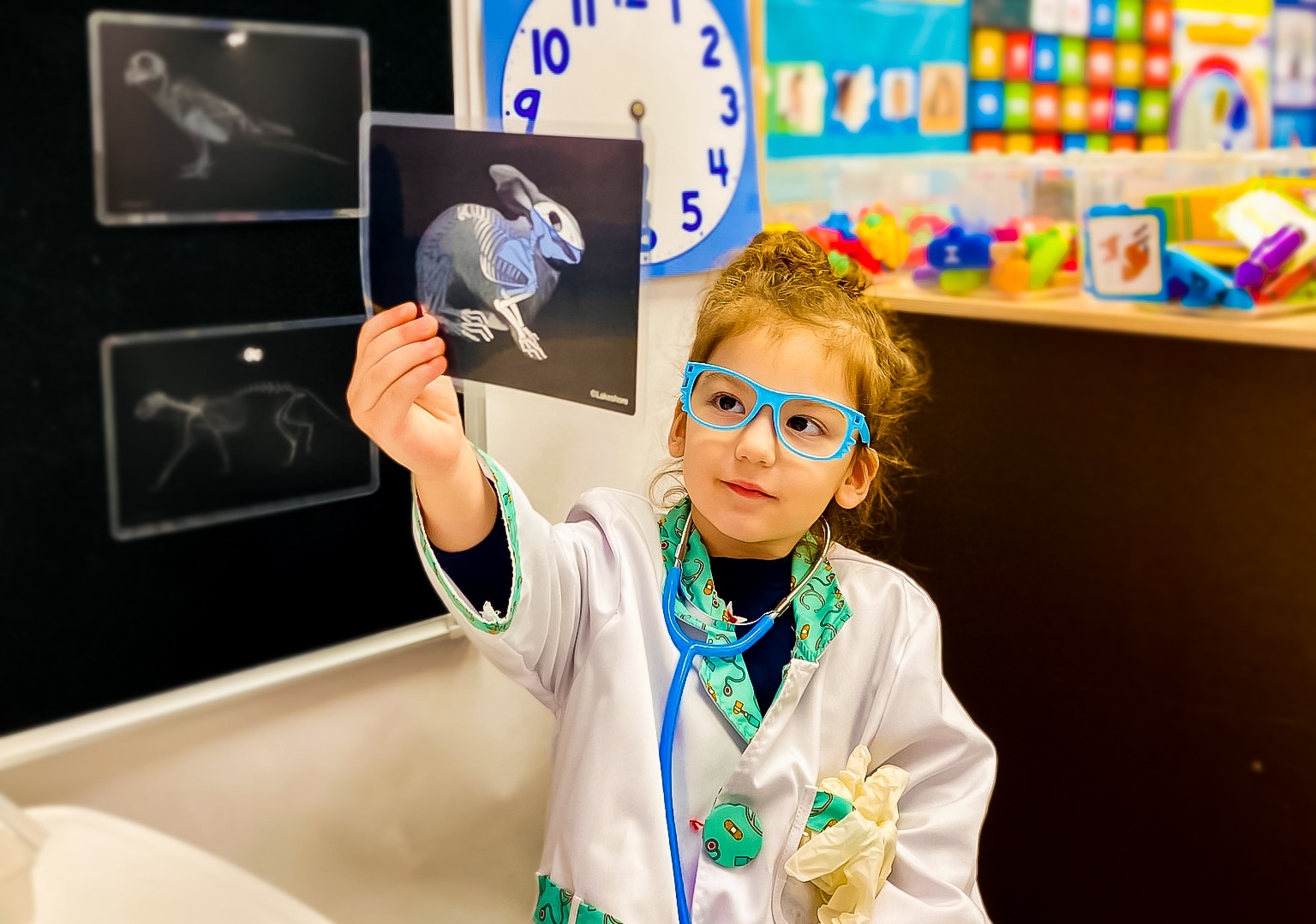 A young girl dressed as a doctor, wearing glasses and a white coat, holding up an X-ray image of a rabbit in a daycare classroom.