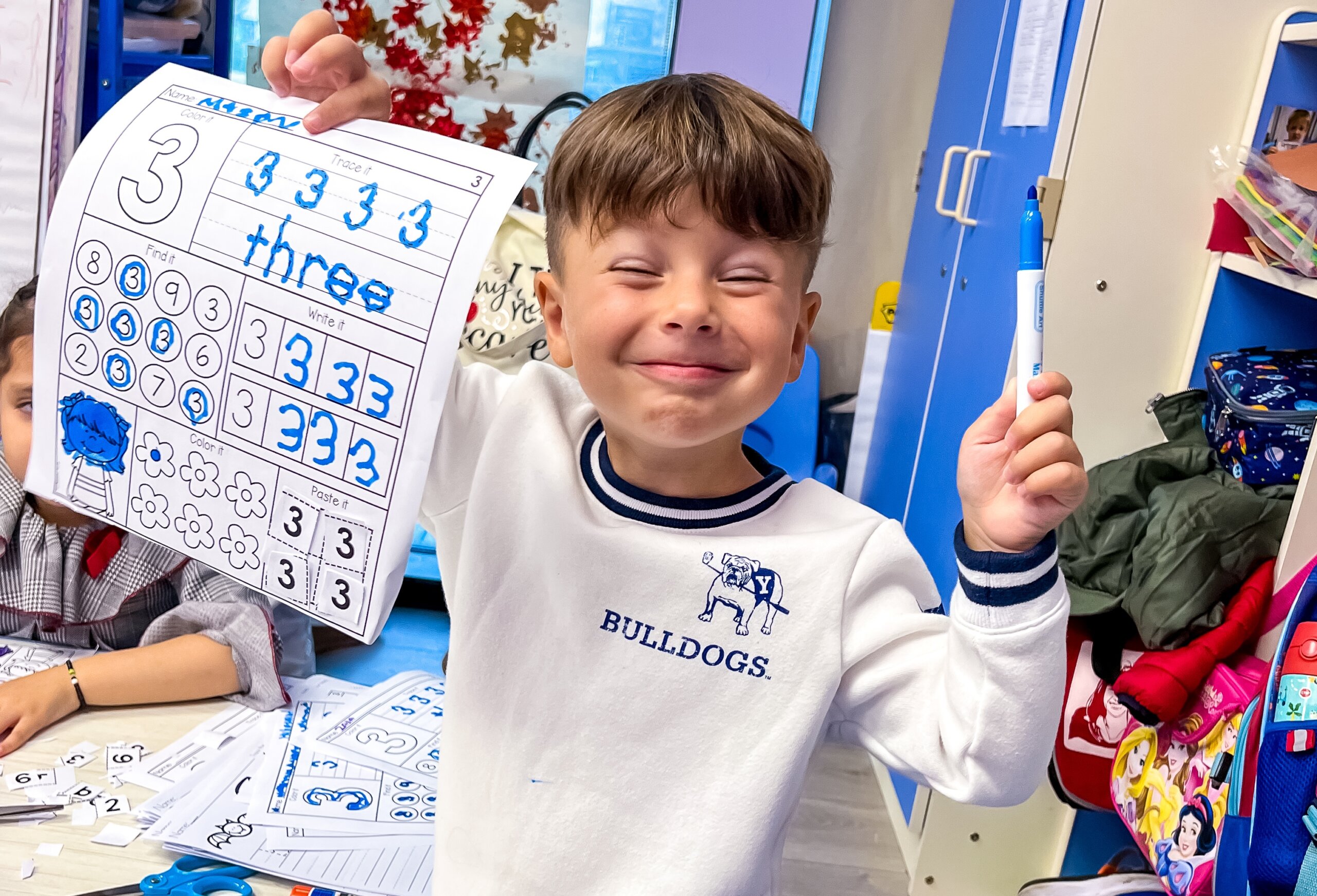 Happy child holding up a worksheet with the number three, showing his completed work in a classroom with educational materials and colorful decorations