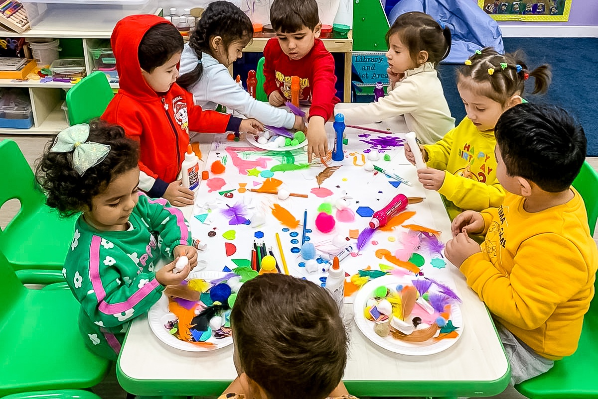 Group of young children sitting around a table, engaged in a colorful craft activity with glue, feathers, pom-poms, and other art supplies in a classroom