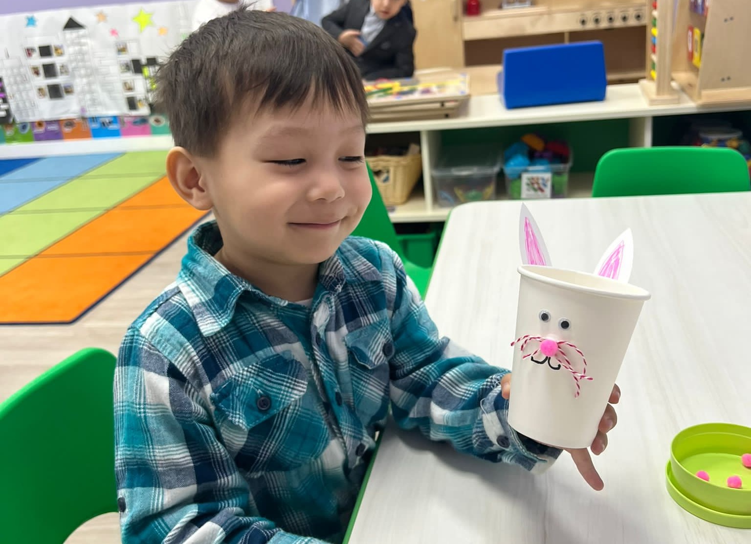 Young boy in a plaid shirt holding a bunny-themed paper cup craft in a daycare classroom