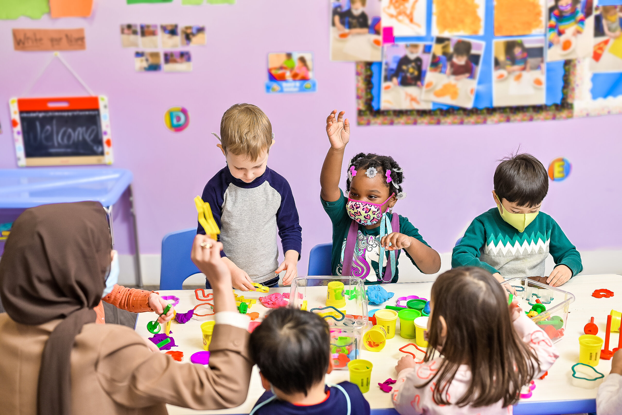 Group of young children wearing face masks sitting around a table doing crafts in a daycare classroom, with one child raising her hand