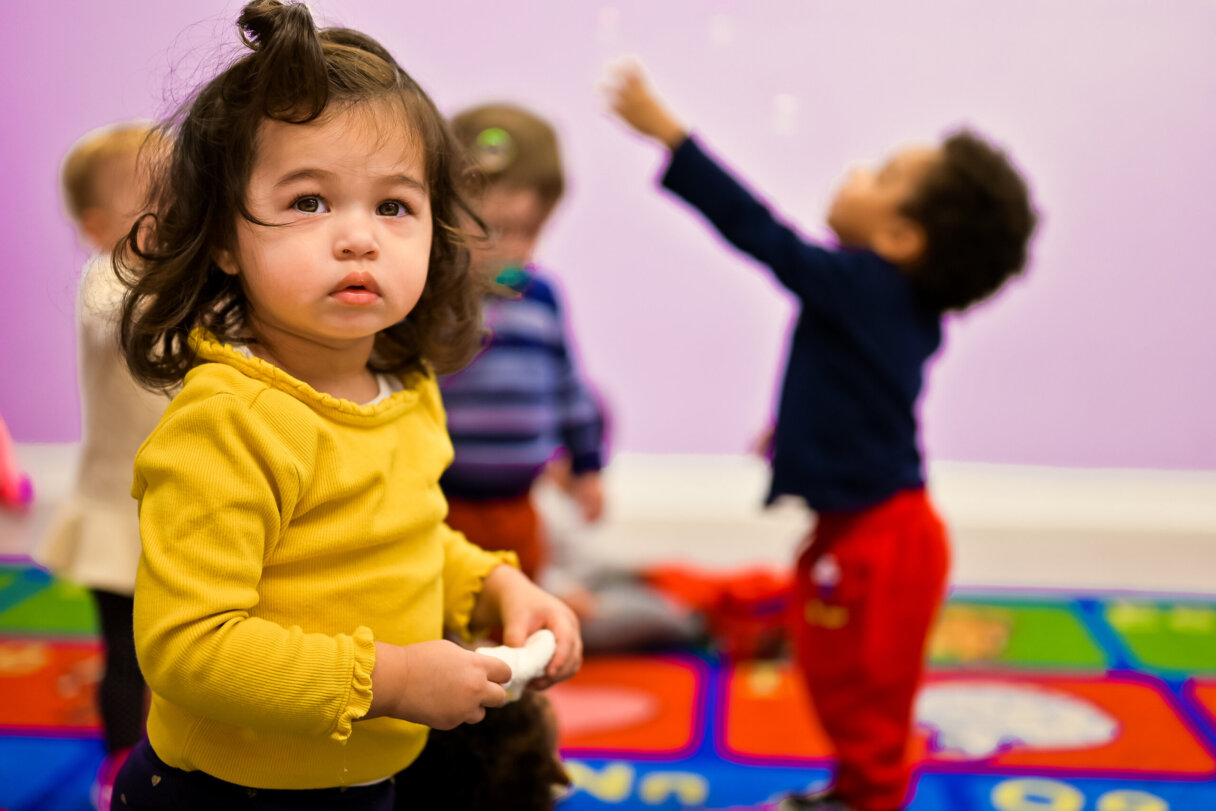 Young girl in a yellow sweater standing and holding a toy, with other children playing in the background on a colorful mat in a classroom.