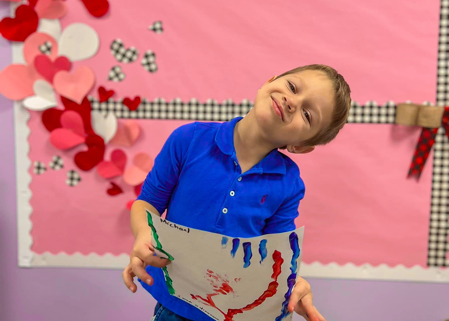 Smiling young boy in a blue shirt holding a colorful painting in a classroom with a pink and heart-decorated bulletin board