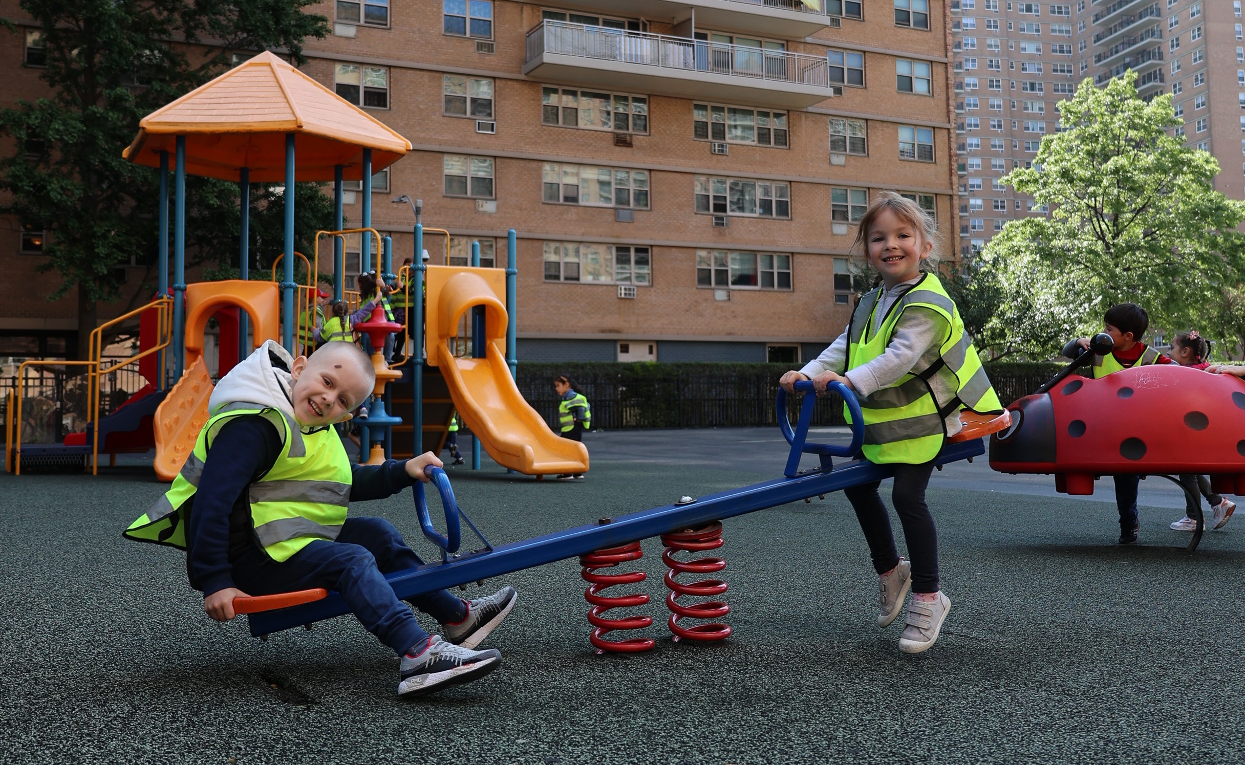 Two children wearing reflective safety vests playing on a seesaw in a playground with slides and climbing structures