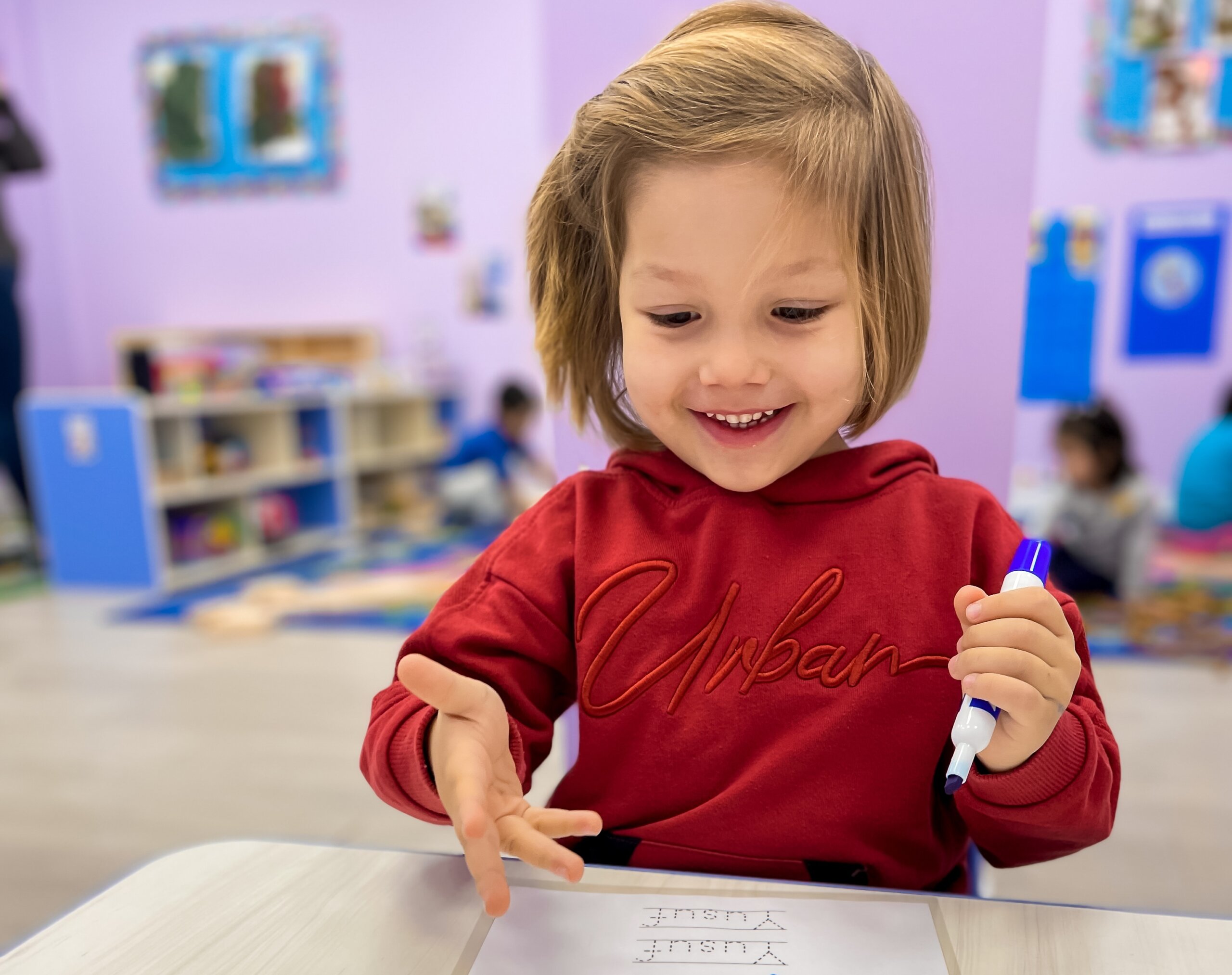 Smiling child in a red hoodie writing on paper with a marker in a colorful classroom setting with other children and toys in the background